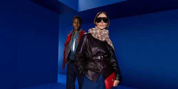 Two fashion models in leather jackets and denim, styled with scarf, sunglasses and red clutch in blue studio.