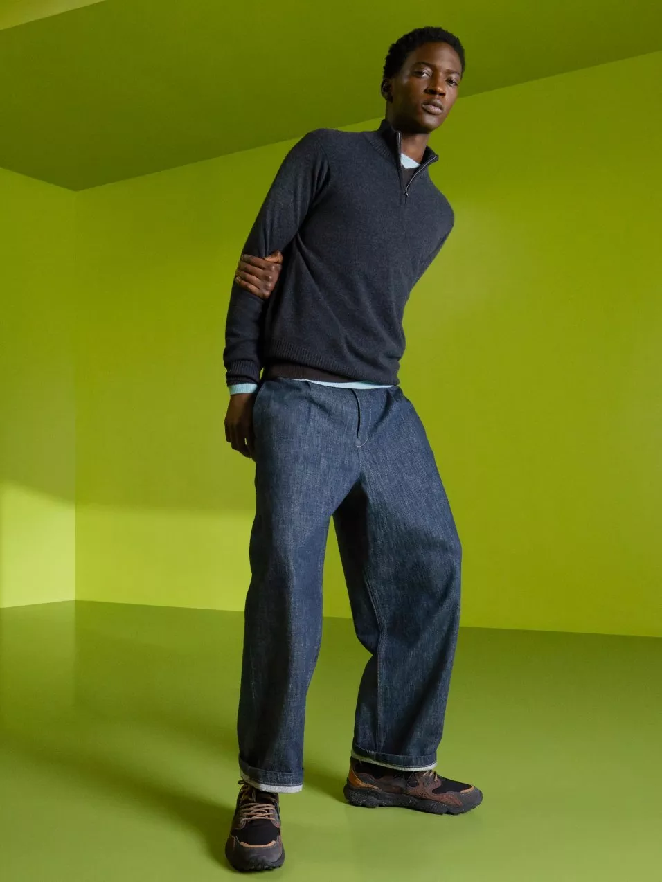 Male fashion model in navy quarter-zip jumper and wide-leg jeans, posing in lime green studio backdrop
