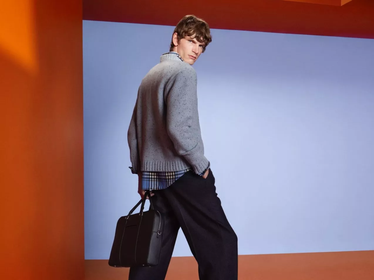 Male model in grey knit jumper and checked shirt holding a black leather briefcase in modern studio setting