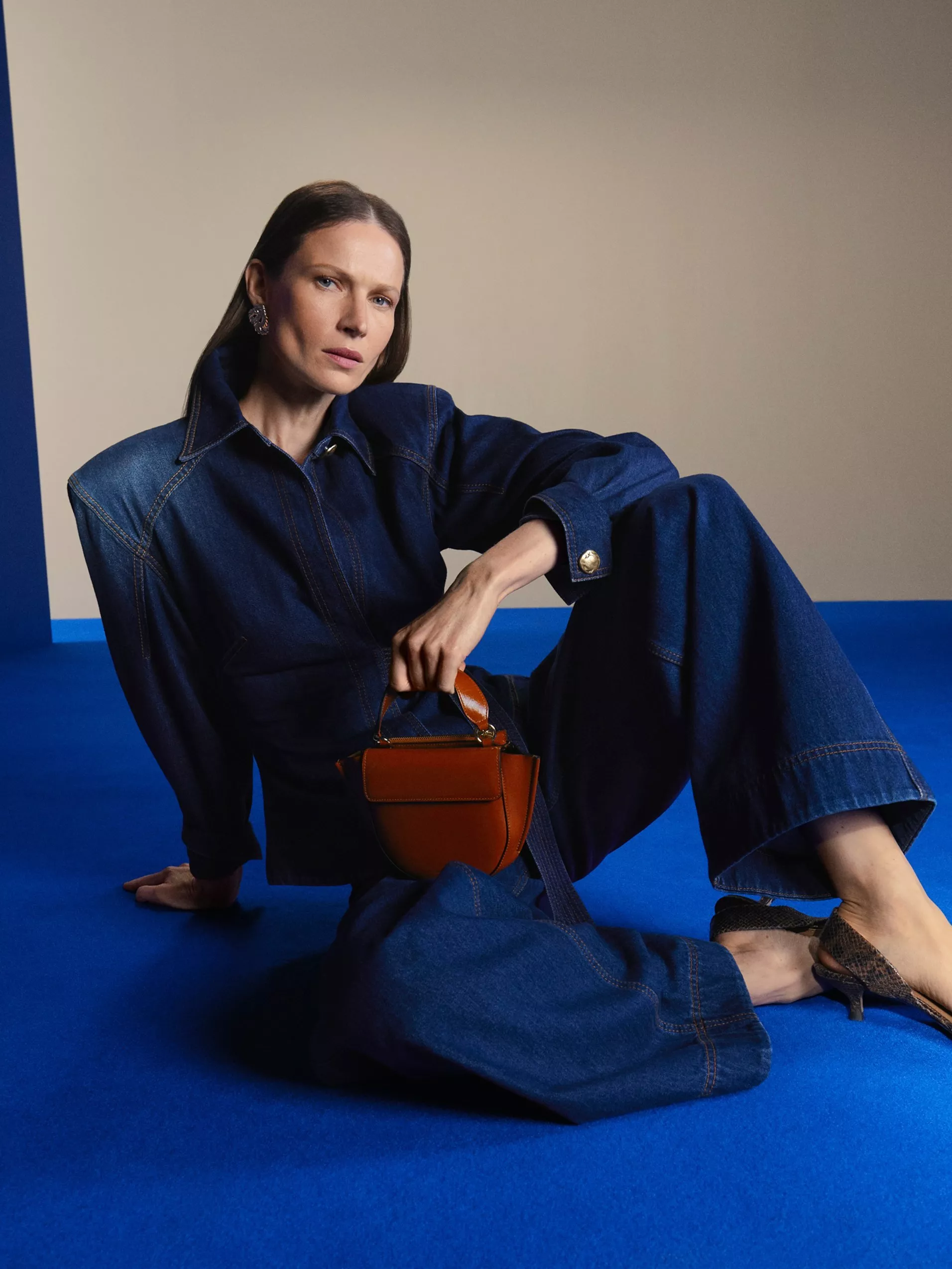 Fashion portrait of woman in denim jumpsuit on blue floor, holding brown handbag, studio backdrop