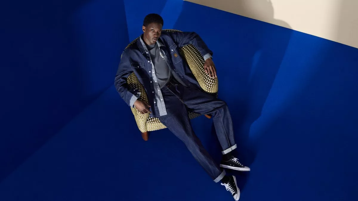 Fashion model in denim jacket and jeans lounging in armchair on blue studio set, casual streetwear look
