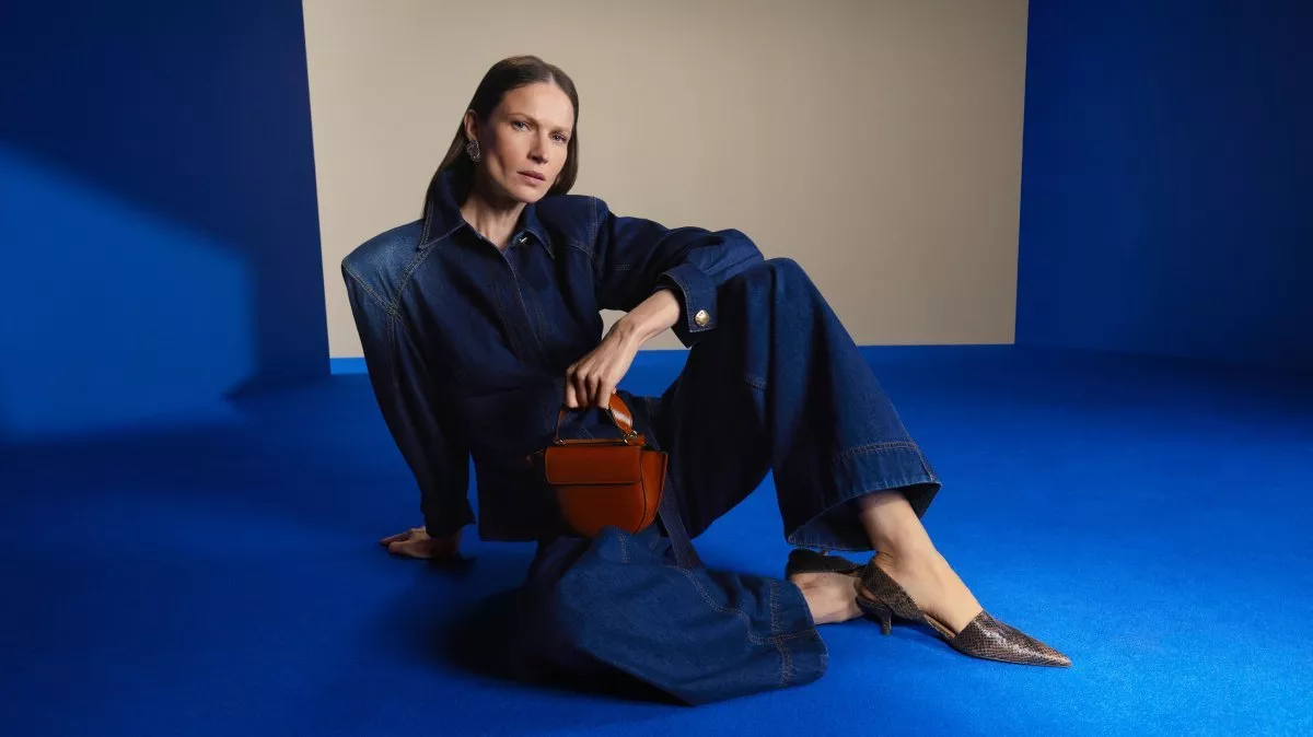 Fashion portrait of woman in dark denim jumpsuit holding brown handbag on blue studio backdrop