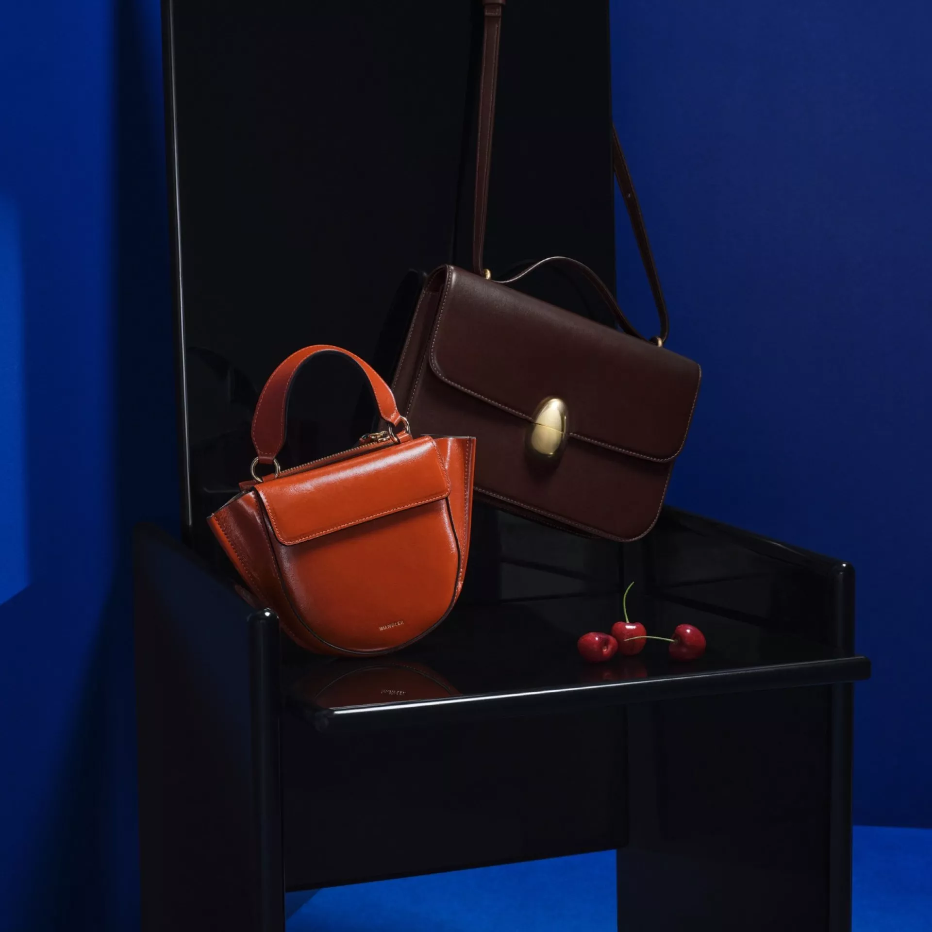 Luxury leather handbags in orange and dark brown on glossy black chair, with cherries on blue studio backdrop