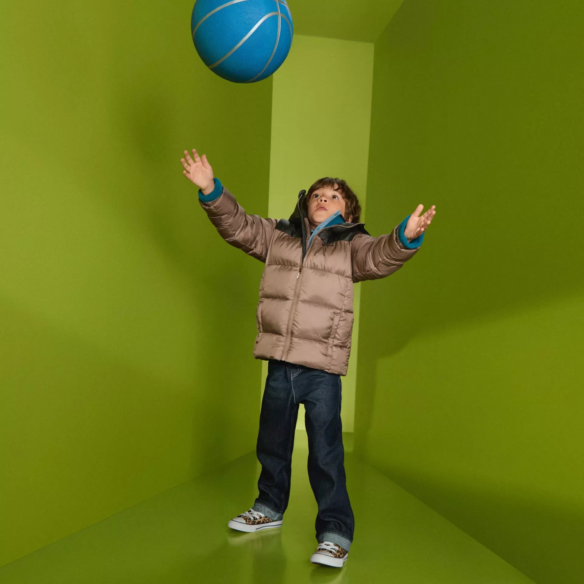 Child in a beige puffer jacket reaching for a blue ball in a green room, kids winter fashion photo