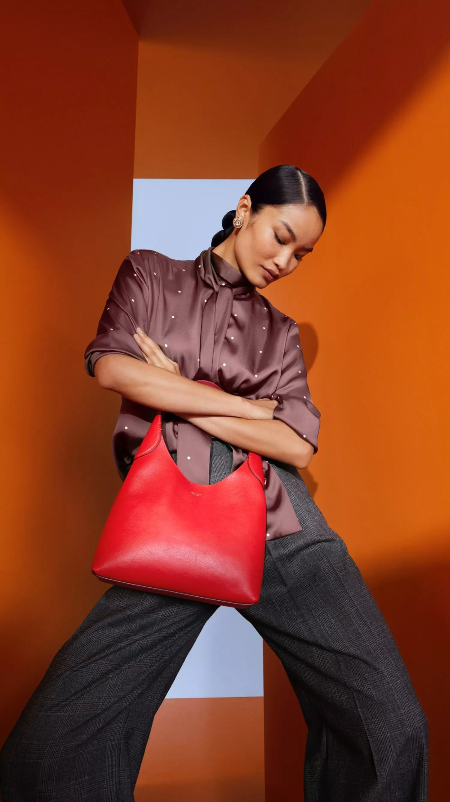 Fashion model posing with bright red leather handbag, brown blouse and grey trousers against orange studio backdrop