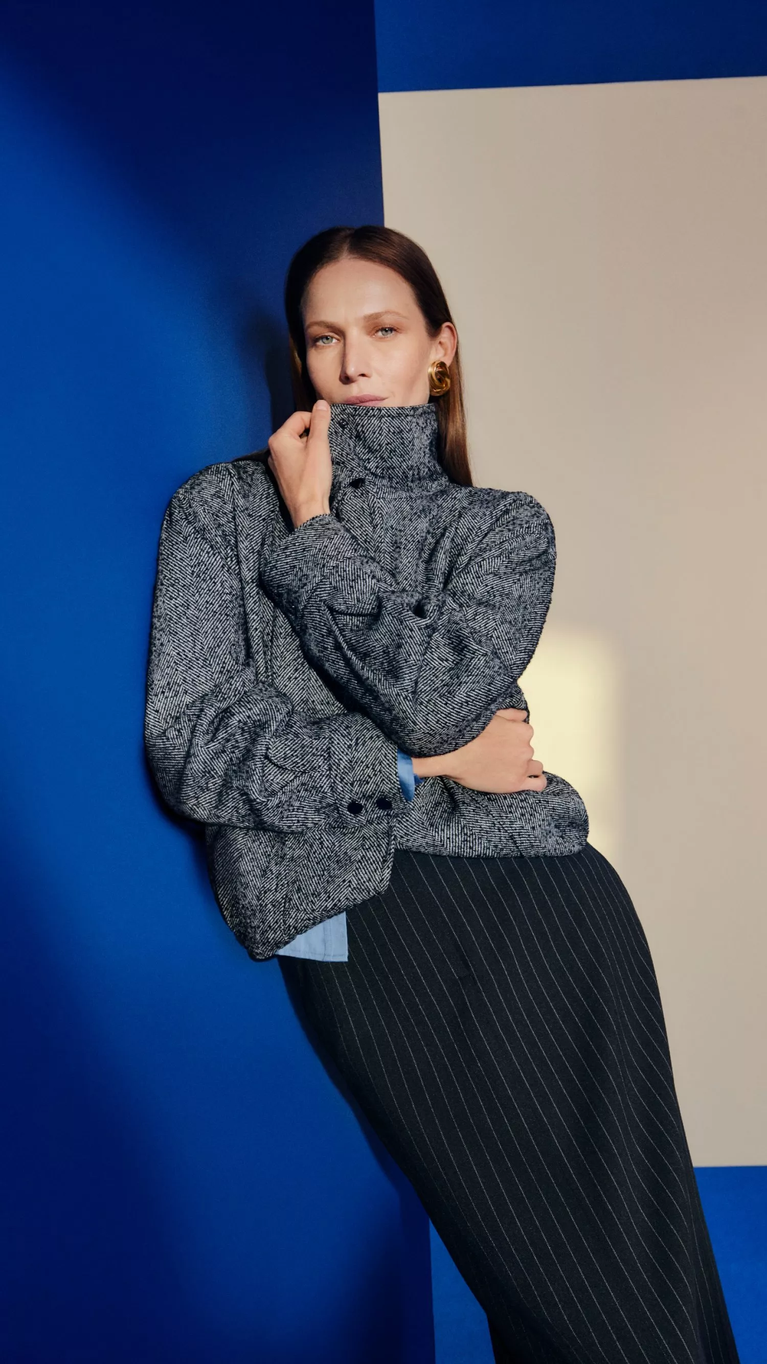Fashion portrait of a woman in a grey tweed jacket and pinstripe trousers against a bold blue wall background