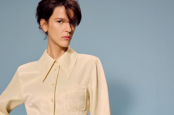 Studio portrait of woman in cream button-up shirt with oversized collar, minimal fashion styling on blue backdrop