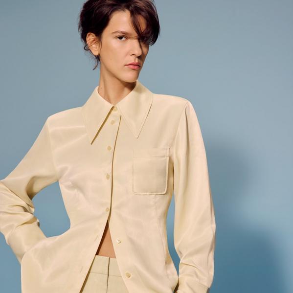 Fashion portrait of a model in a cream satin button-up shirt with oversized collar, posed against a blue studio backdrop
