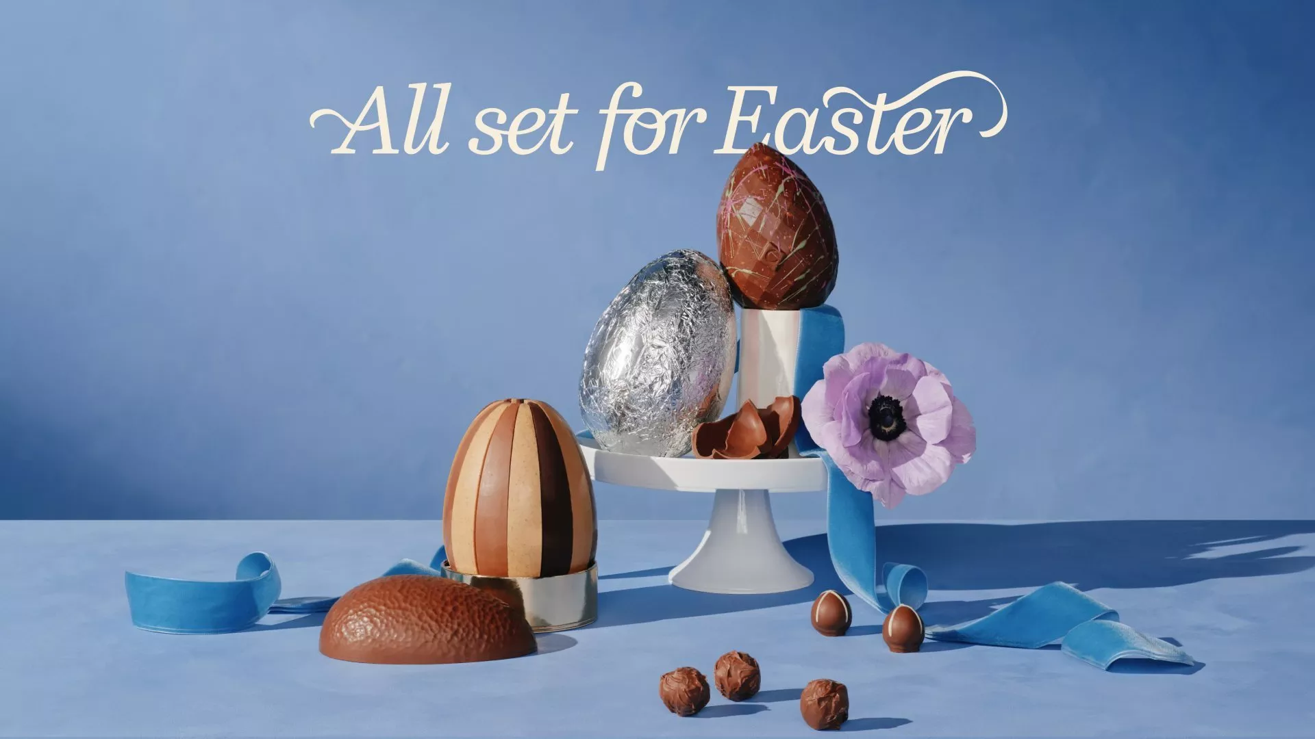 Elegant Easter display with decorative chocolate eggs, a purple flower, and blue ribbon against a blue backdrop.