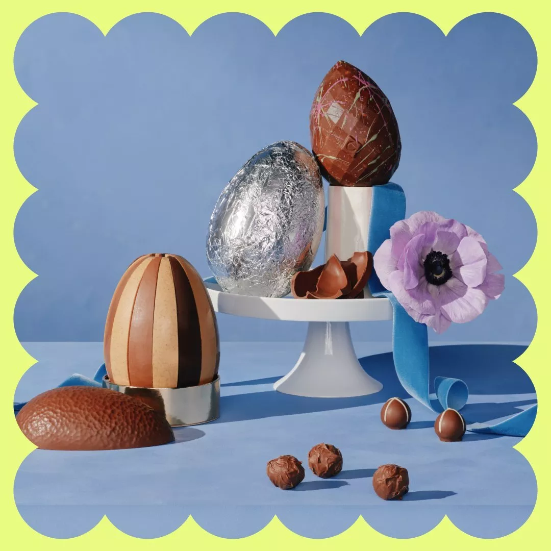 Decorative chocolate eggs on a stand with a purple flower, set against a blue background with a scalloped border.