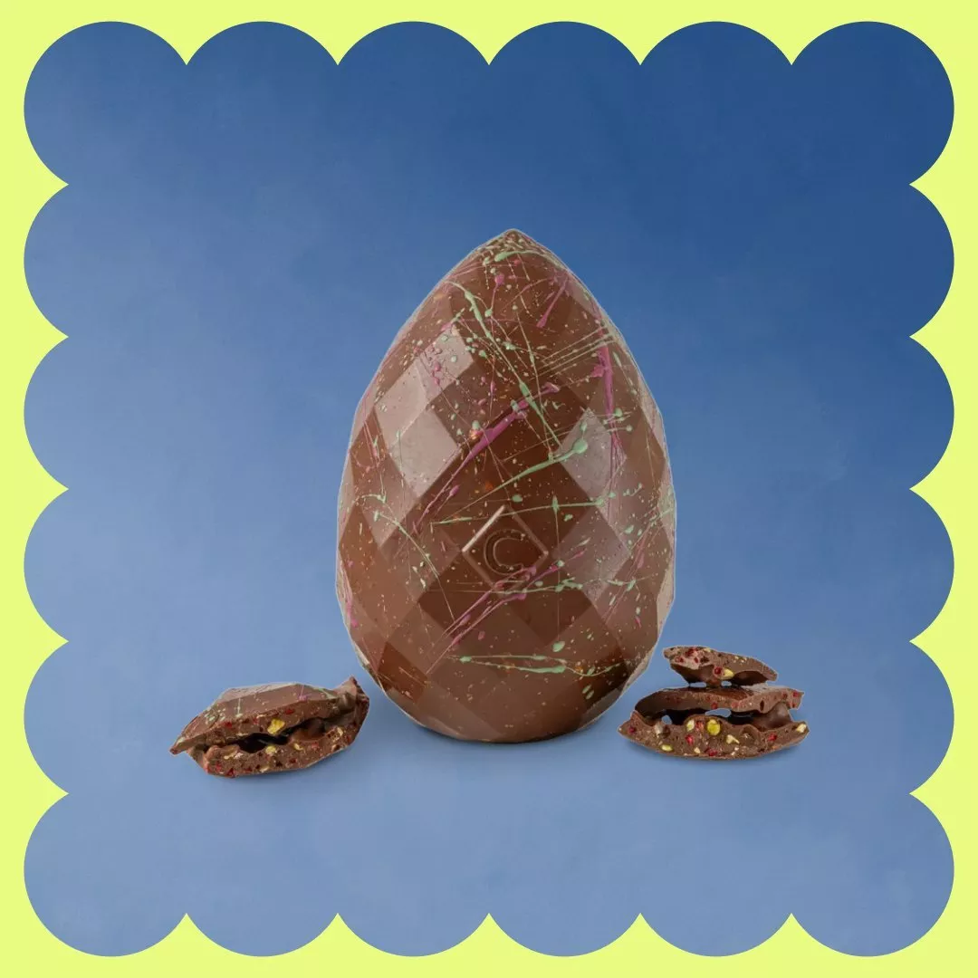 Geometric chocolate egg with colourful splatter design, accompanied by broken chocolate pieces, on a blue background.