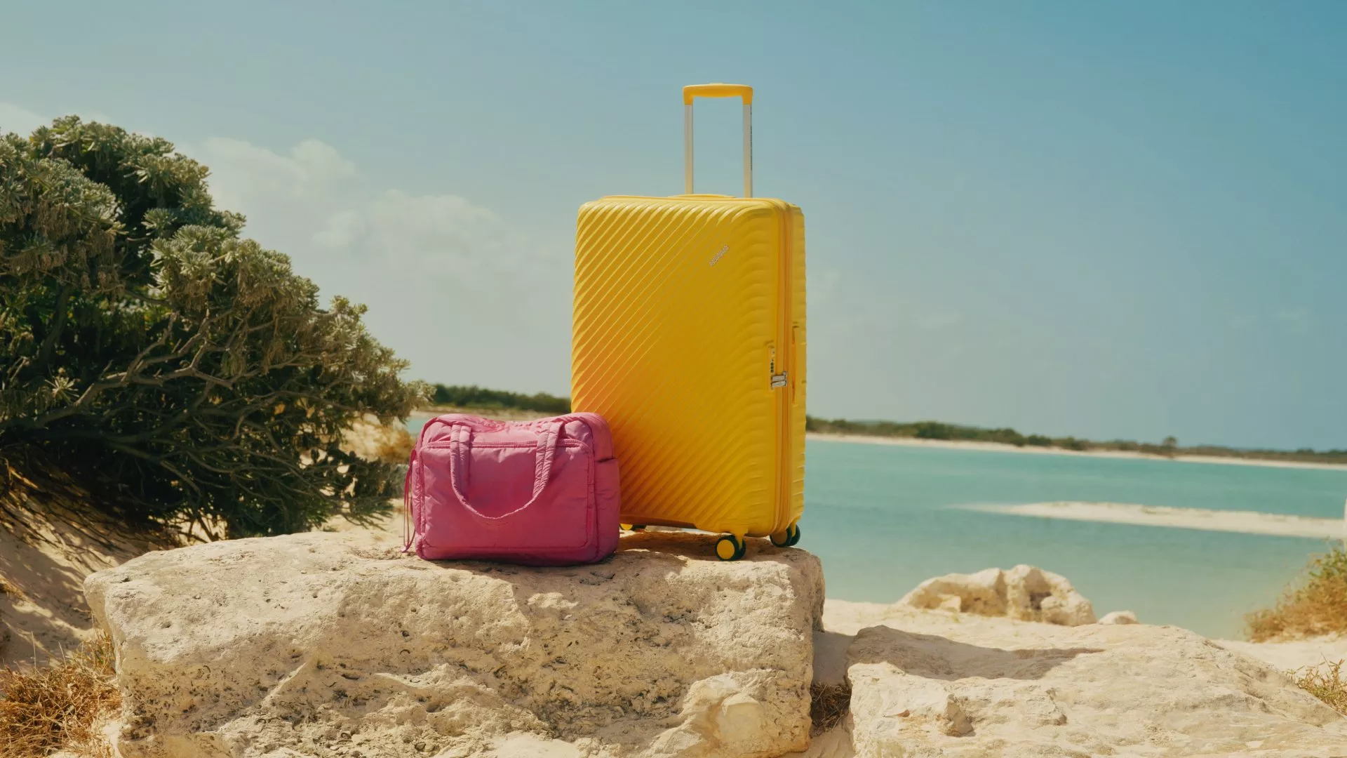 Yellow hard-shell suitcase and pink travel bag on rocks by a sunny beach, summer holiday luggage scene