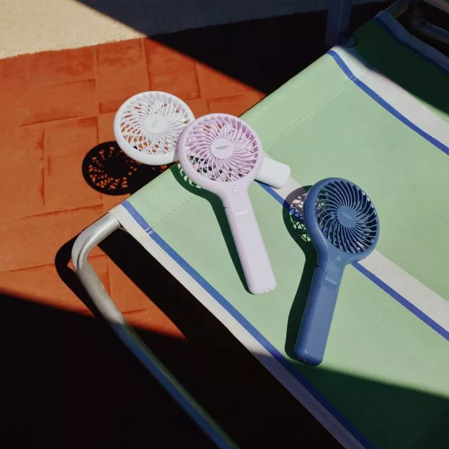Three portable handheld mini fans on a green striped sun lounger on patio tiles, summer travel cooling gadgets