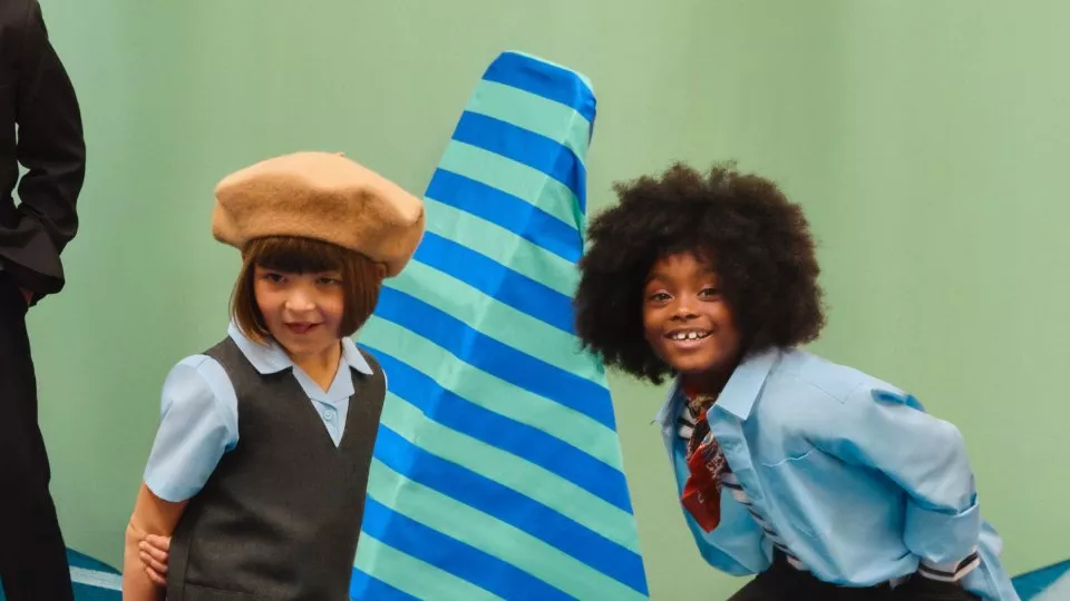 Two children in school uniforms posing in a studio, with a large blue striped prop between them