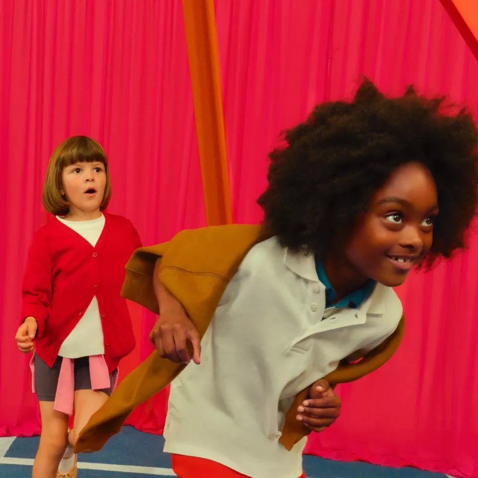 Two children playing together indoors, one running in a white polo, bright pink drapes backdrop, joyful scene