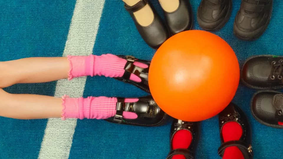 Top view of schoolchildren’s feet around an orange playground ball on a blue carpeted floor indoors