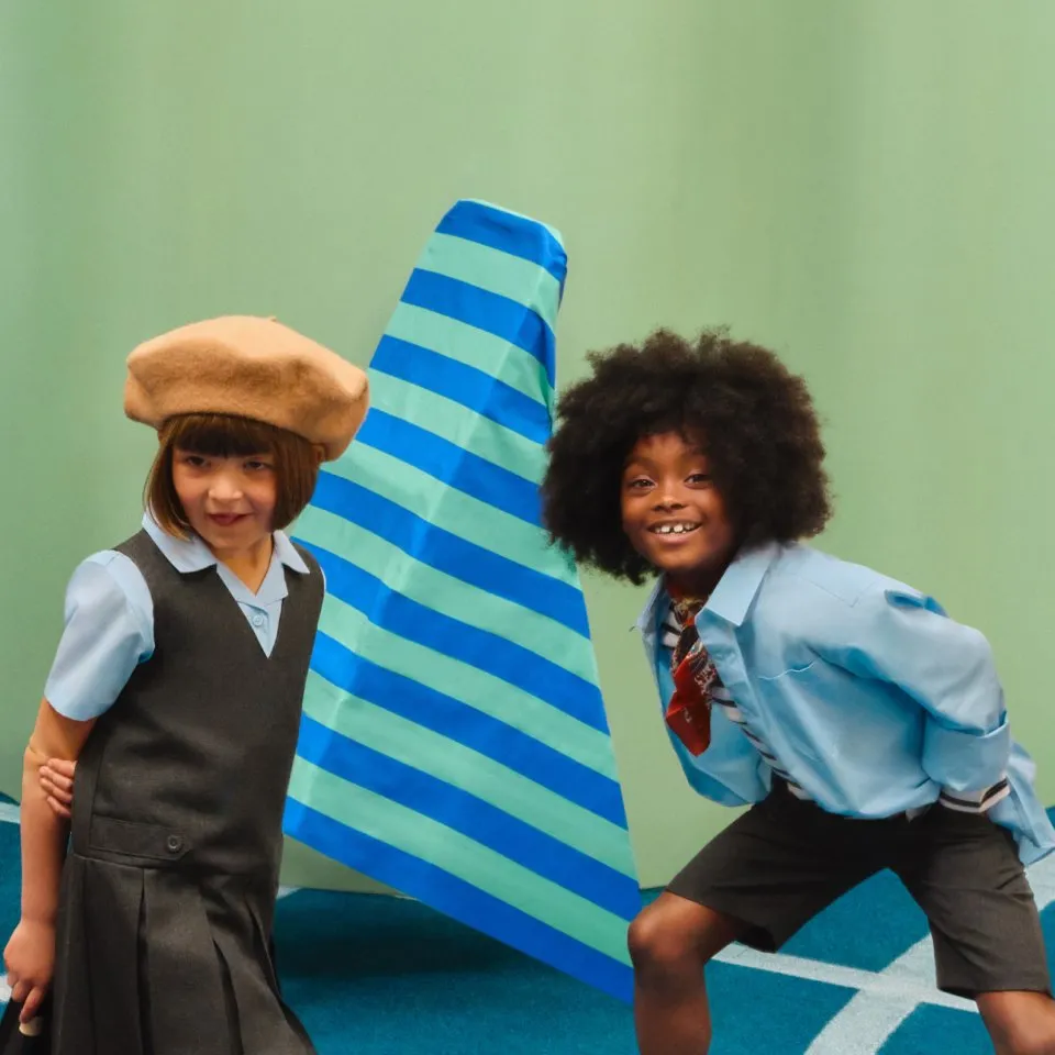 Two children in retro school uniforms smiling, posing beside a blue striped cone on a green studio backdrop