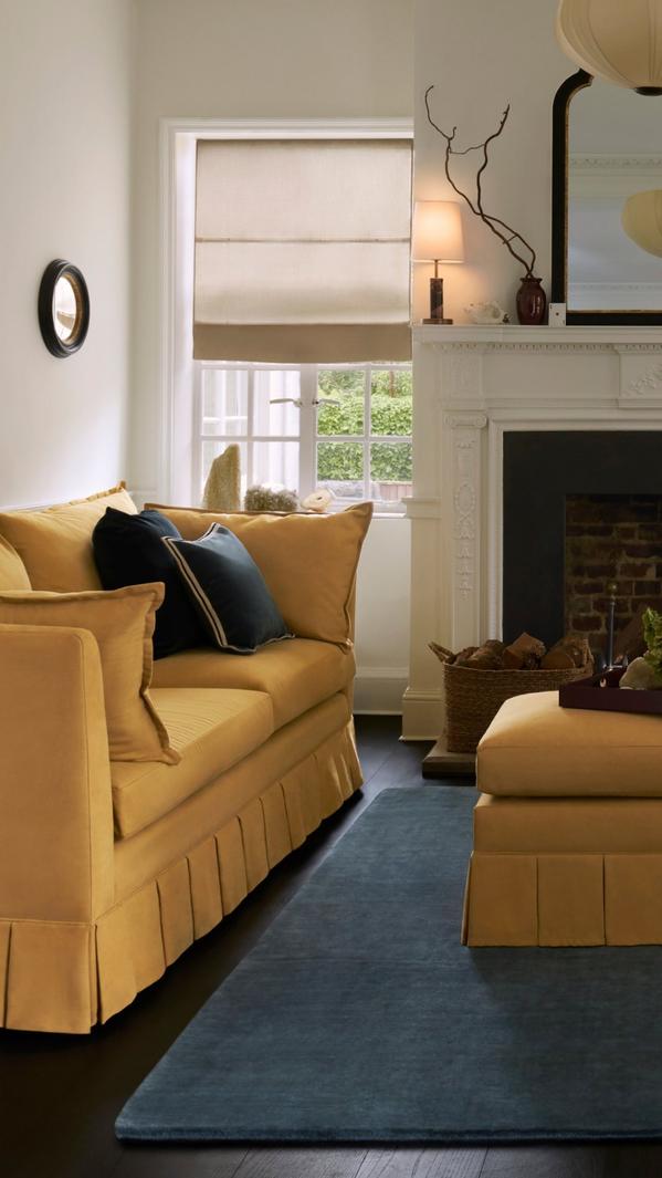 Cosy living room with mustard yellow sofa, blue rug, Roman blind window and white fireplace mantel decor