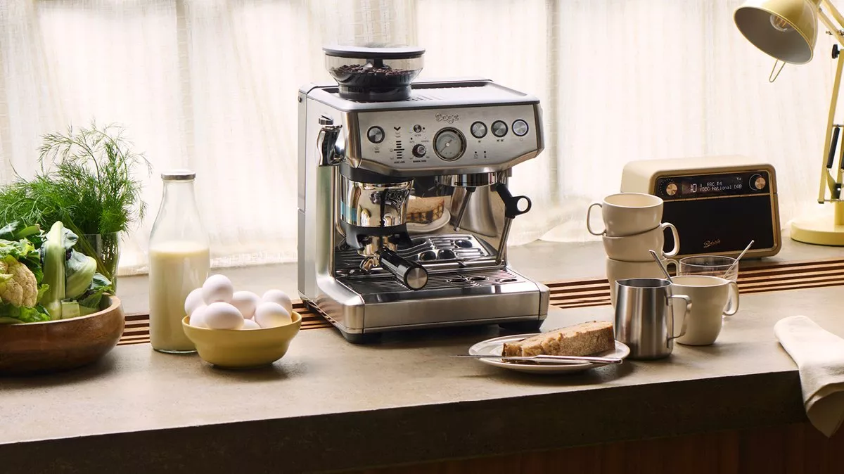 Stainless steel espresso machine on kitchen counter with milk bottle, eggs, mugs, and breakfast toast in sunlight