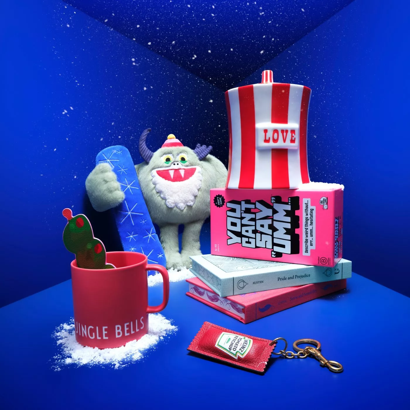 Festive still life with yeti plush, snowboard, LOVE ornament, books stack, cactus mug and keyring on blue backdrop