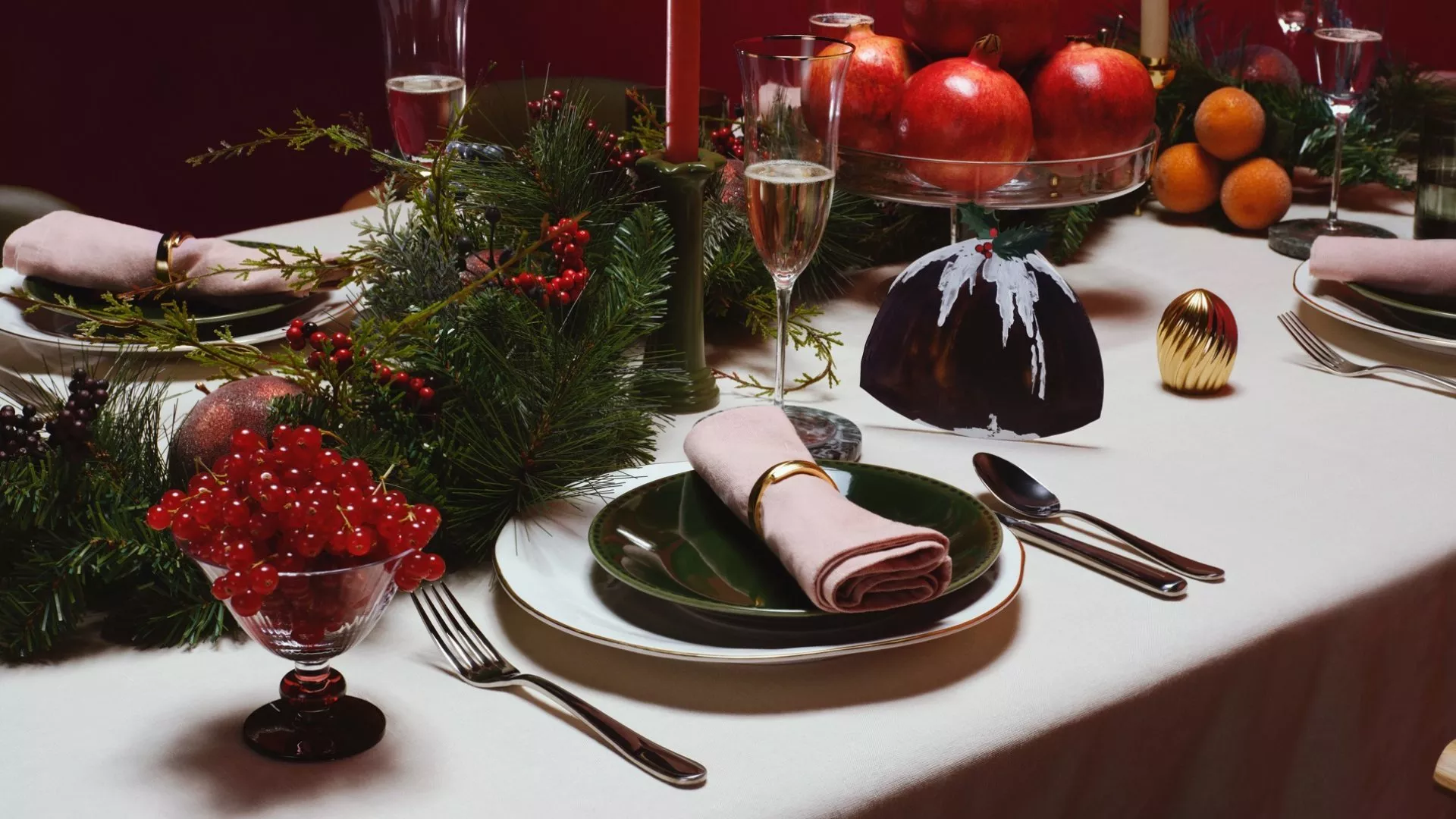 Elegant Christmas dinner table setting with festive greenery, red berries, candles, champagne and fruit centrepiece
