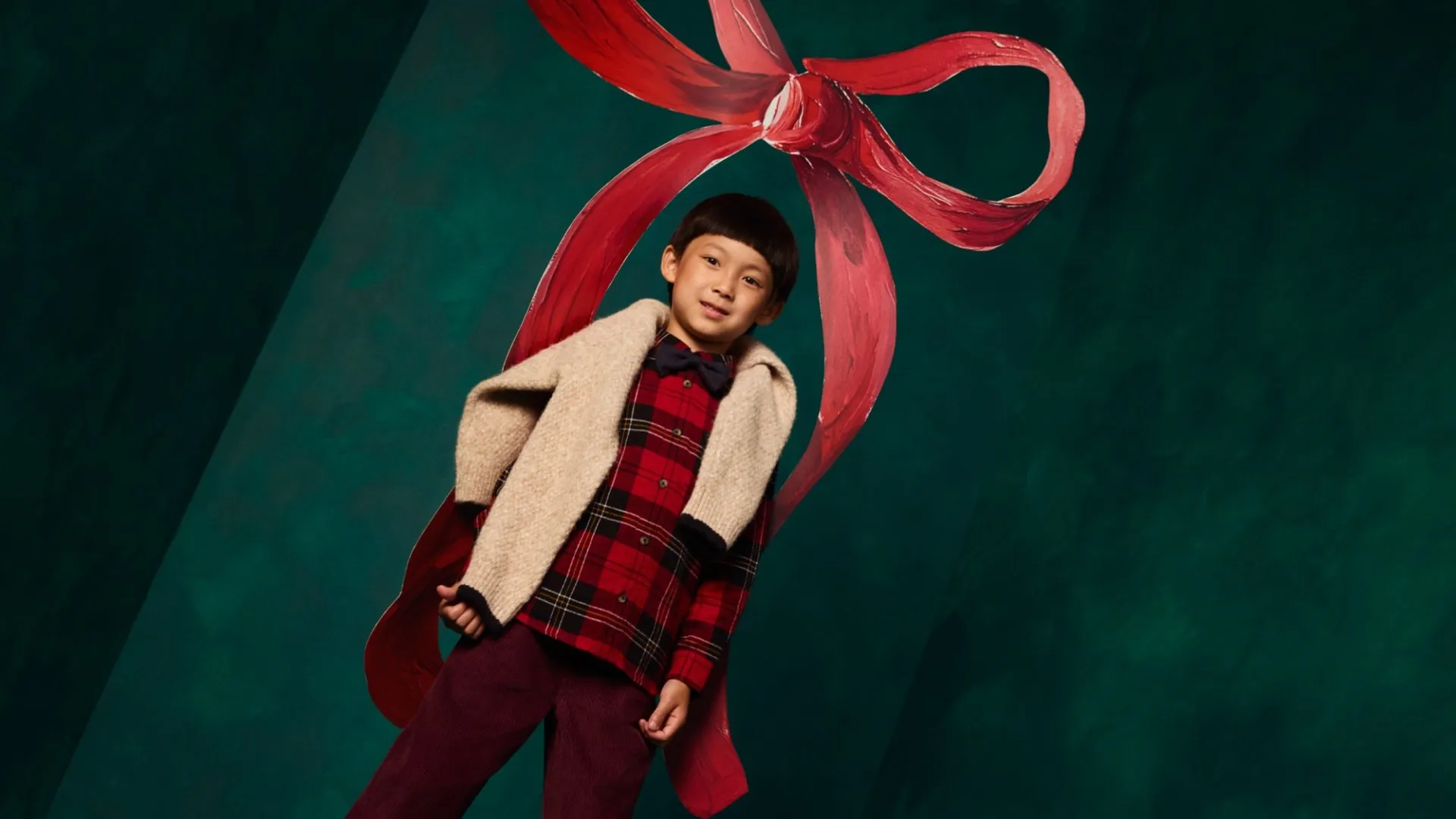 Young boy in festive red tartan shirt and scarf beneath a large red ribbon bow on green studio backdrop