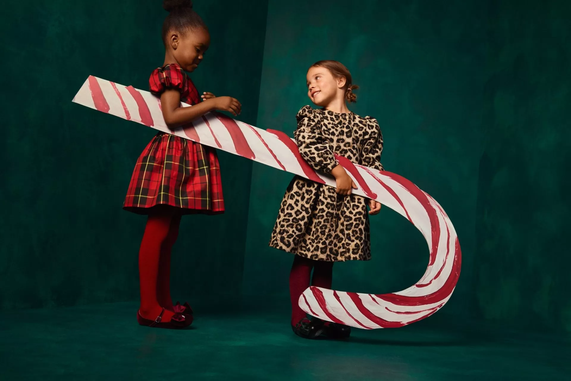 Two girls in festive party dresses holding a giant candy cane prop in a studio Christmas fashion shoot