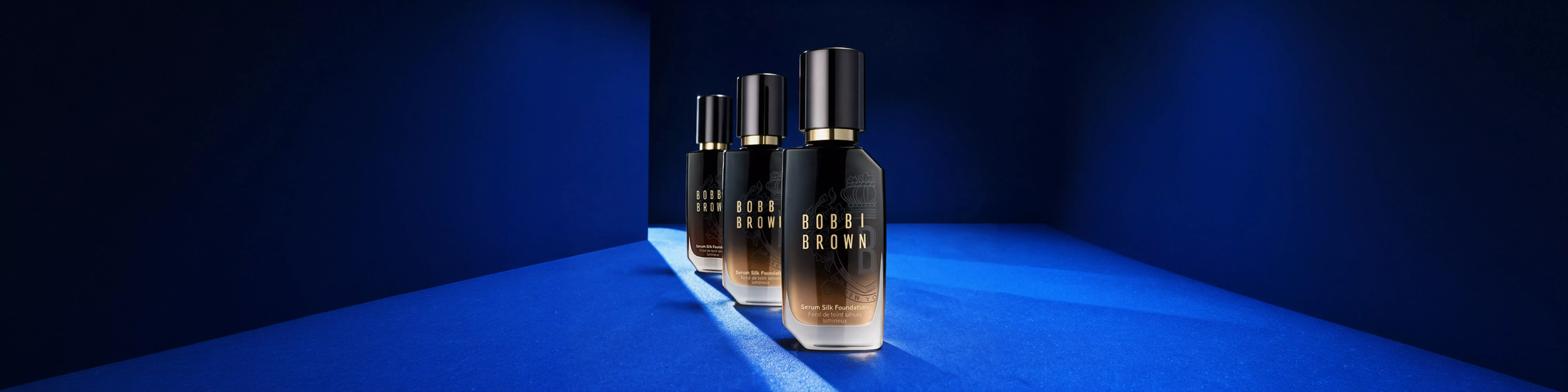 Bobbi Brown Serum Silk Foundation bottles on blue backdrop, luxury makeup product photography with dramatic lighting