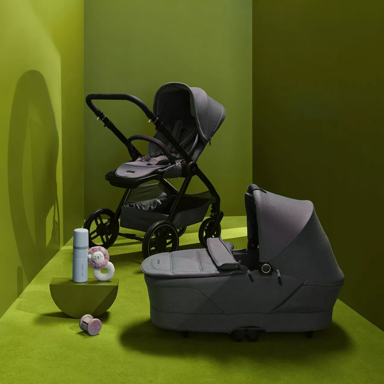 Grey pram and carrycot travel system with baby bottle and toy, photographed in a modern green studio setting