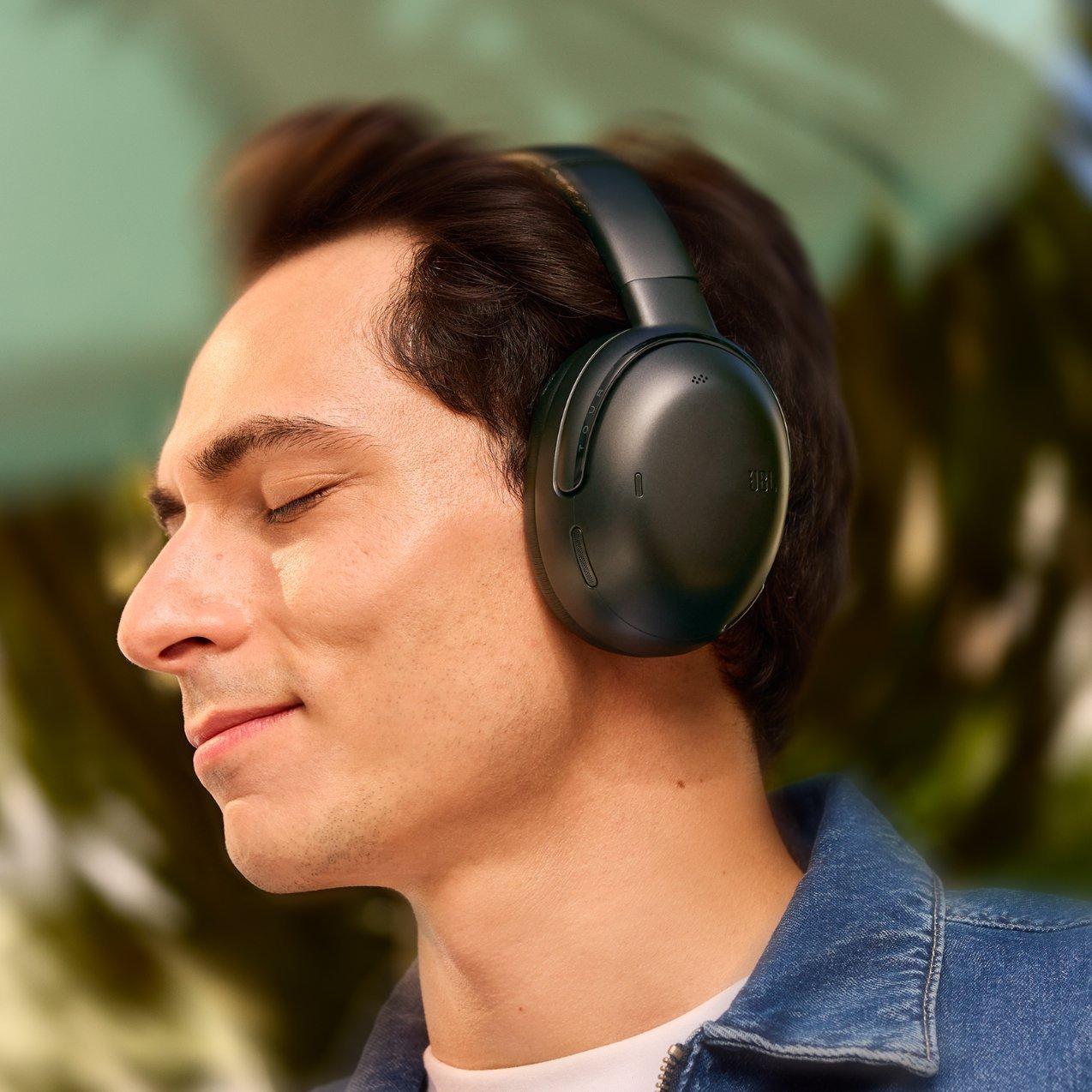 A man with his eyes closed and a contented smile, wearing sleek black over-ear JBL headphones while listening to music outdoors.