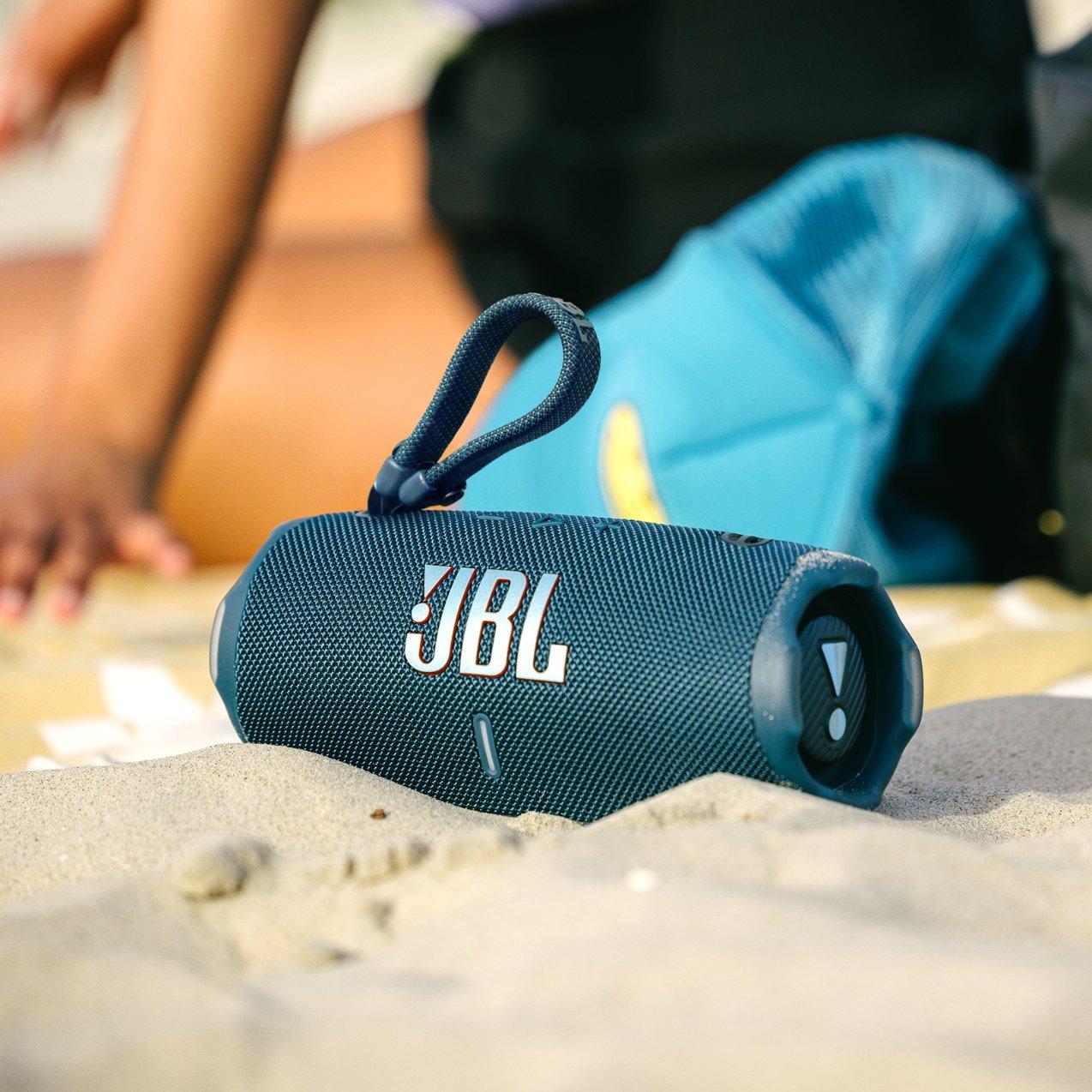 A blue portable JBL Bluetooth speaker resting on a sandy beach next to a blue mesh hat and bag.