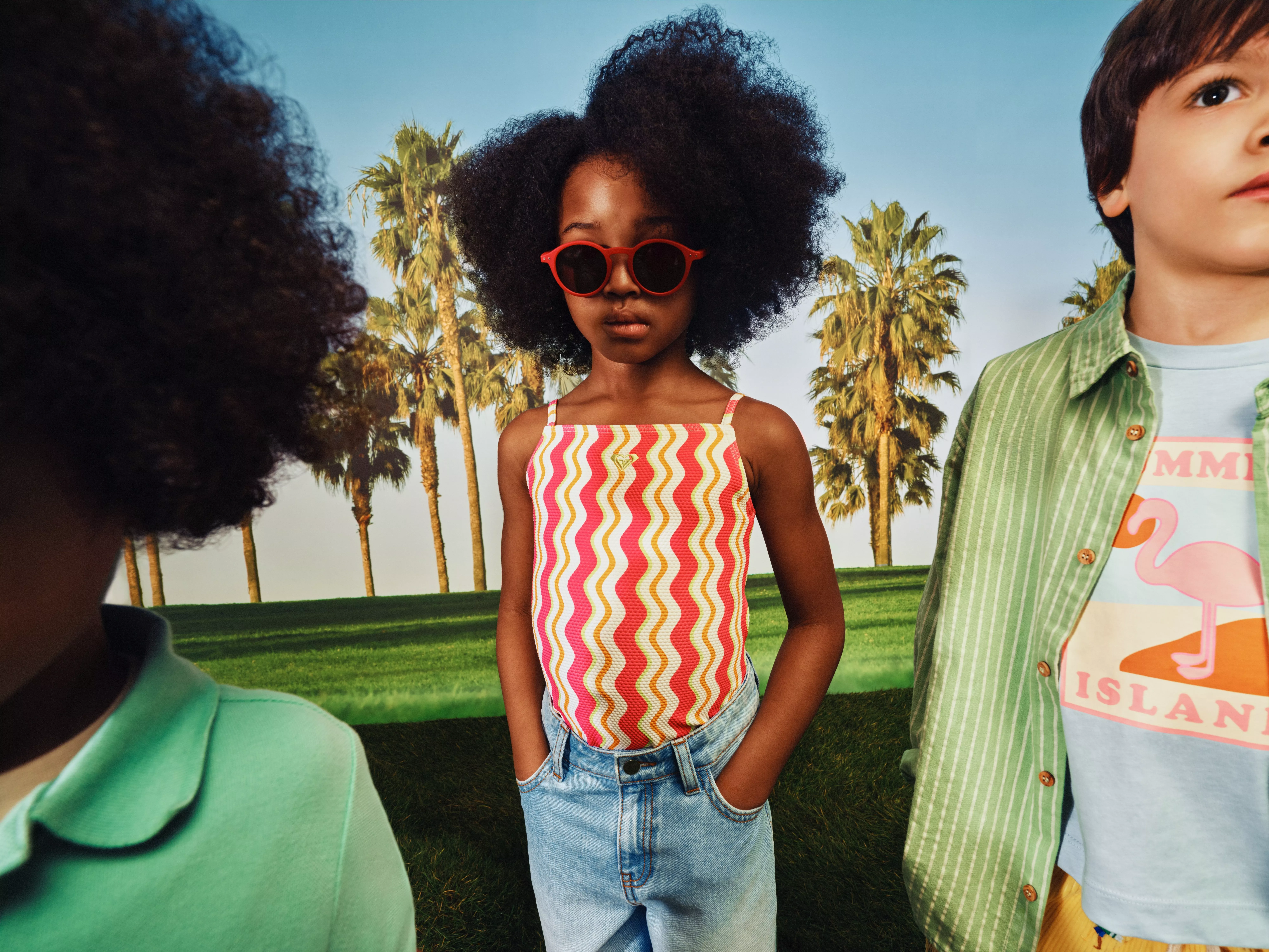 Stylish children in summer outfits, girl in red sunglasses, standing on grass with palm trees in background