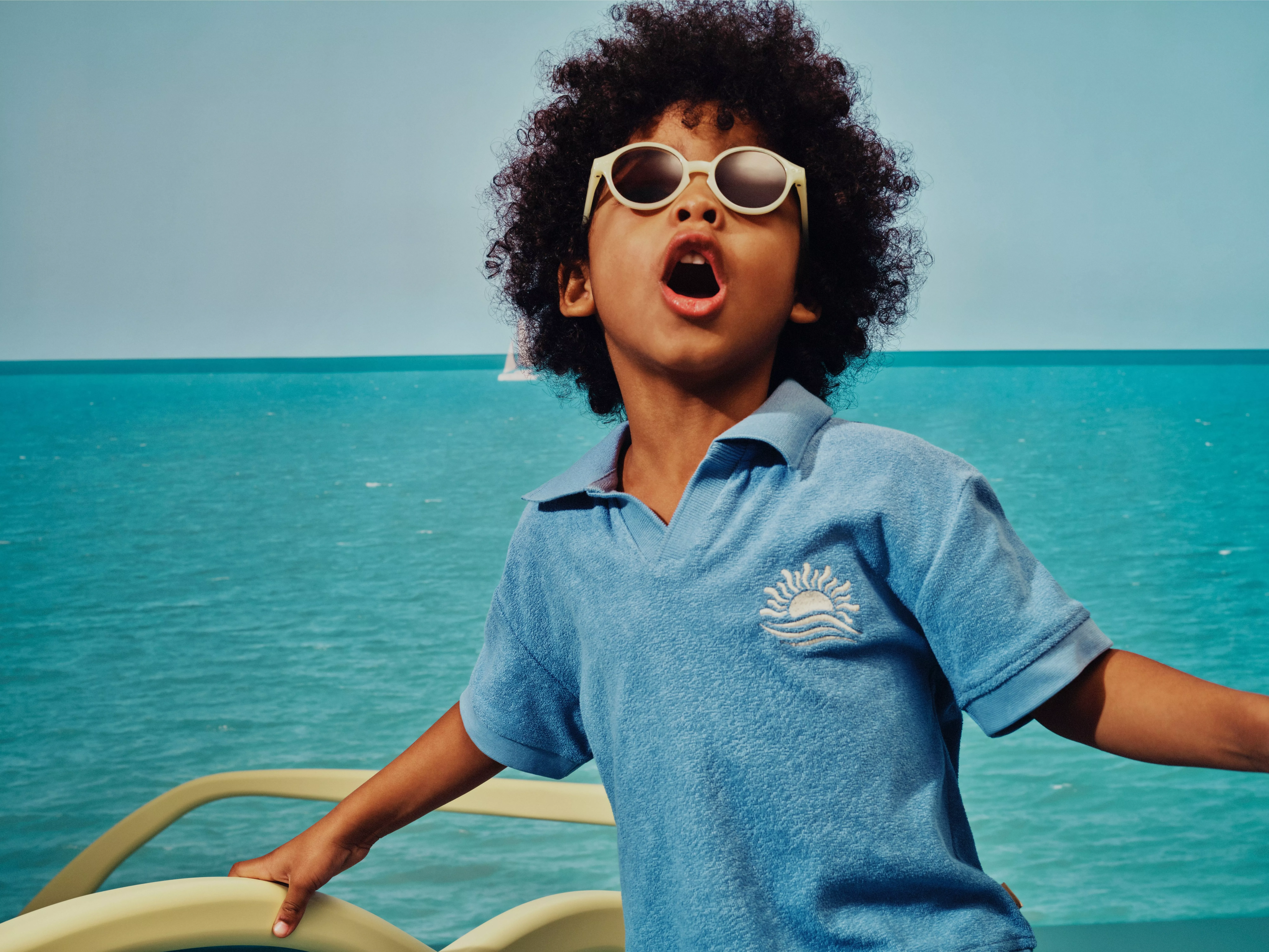 Child in sunglasses on a boat, wearing blue polo shirt, with turquoise sea and sailboat in background