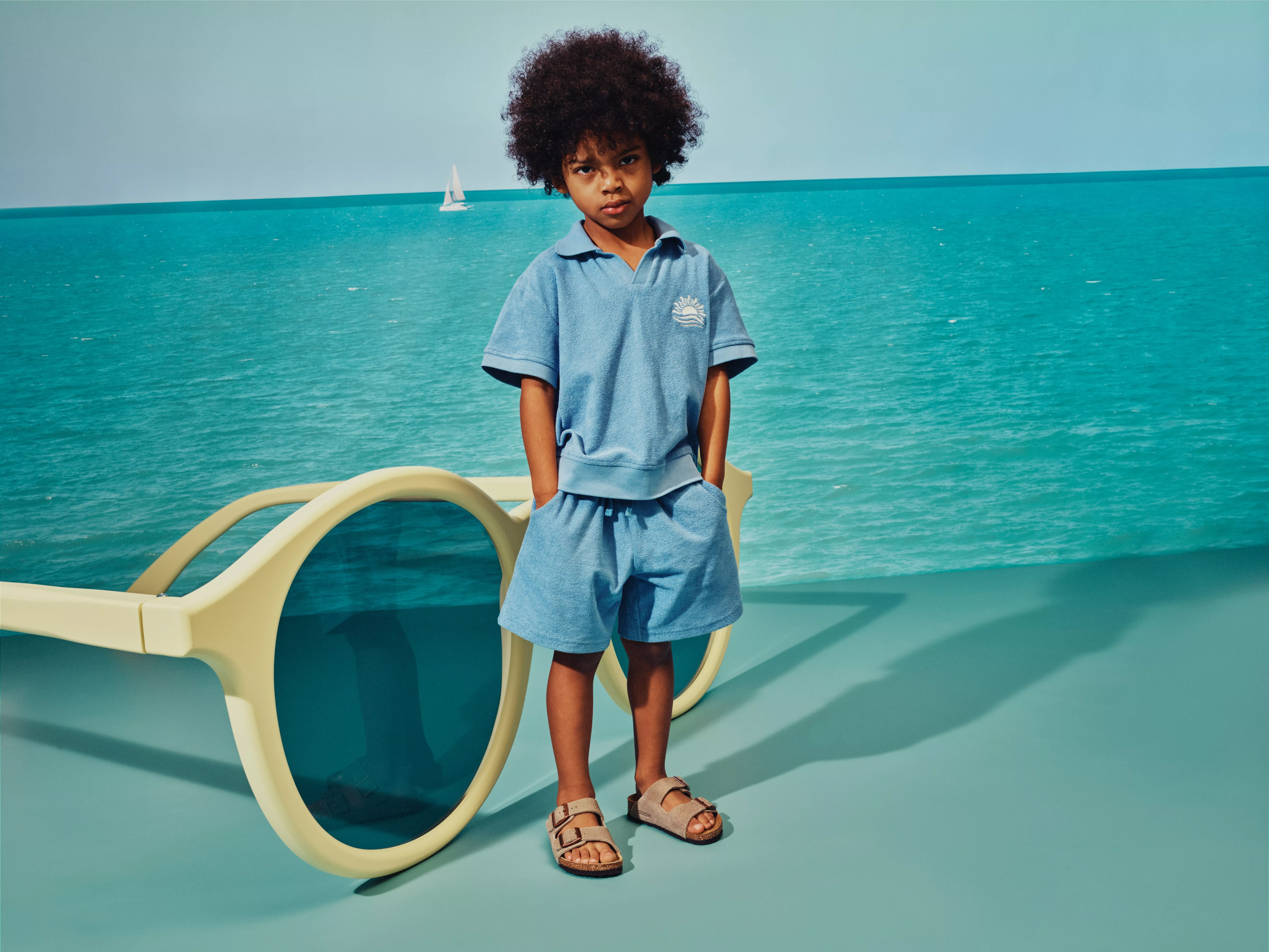Child wearing a blue towelling polo and shorts by the sea, beside oversized sunglasses and a distant sailboat.