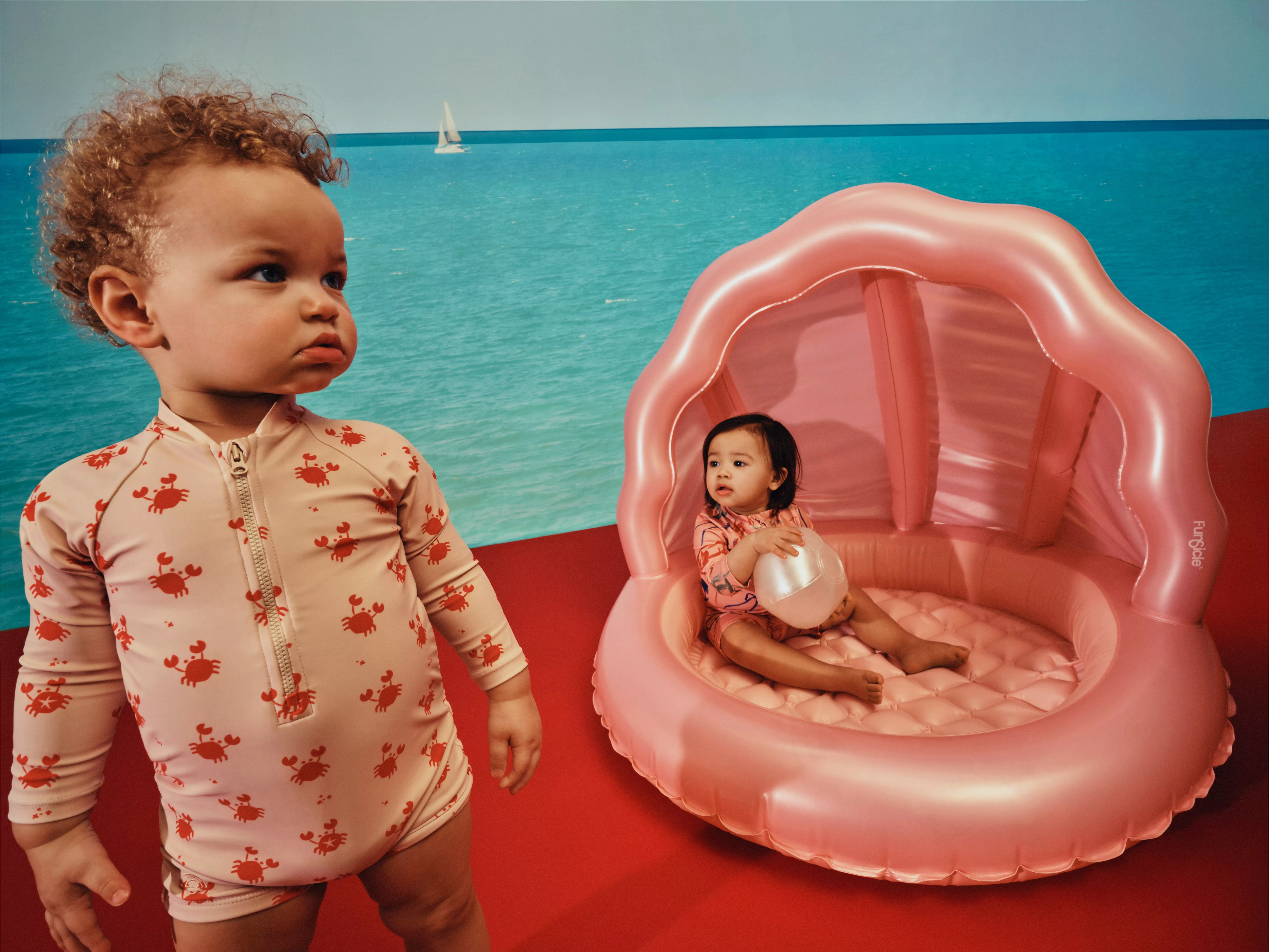 Two toddlers in crab-print swimwear by a pink inflatable baby pool, with a sea mural backdrop indoors