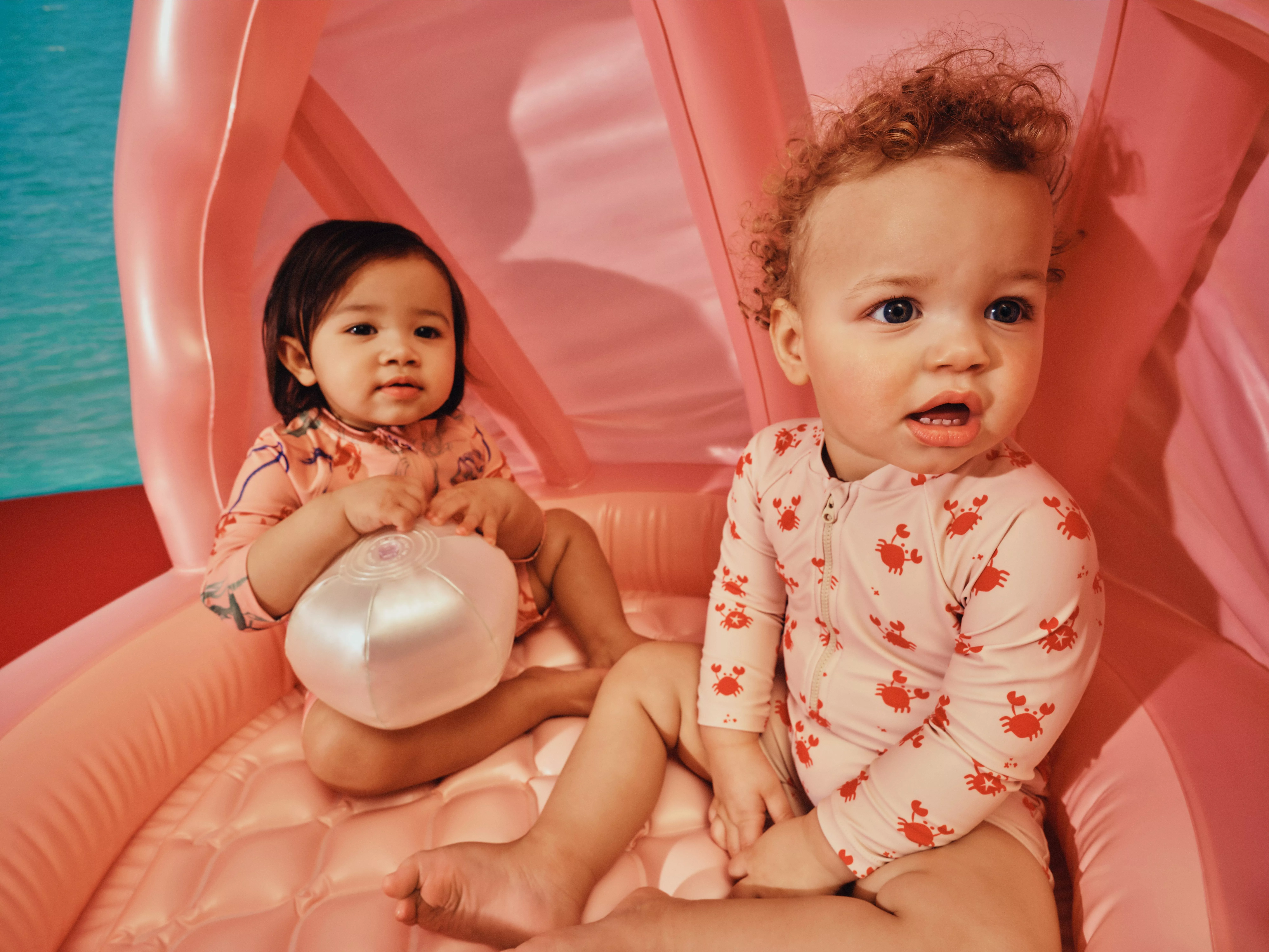 Two babies in pink crab-print swimsuits sitting on a pink inflatable pool float, holding a beach ball outdoors