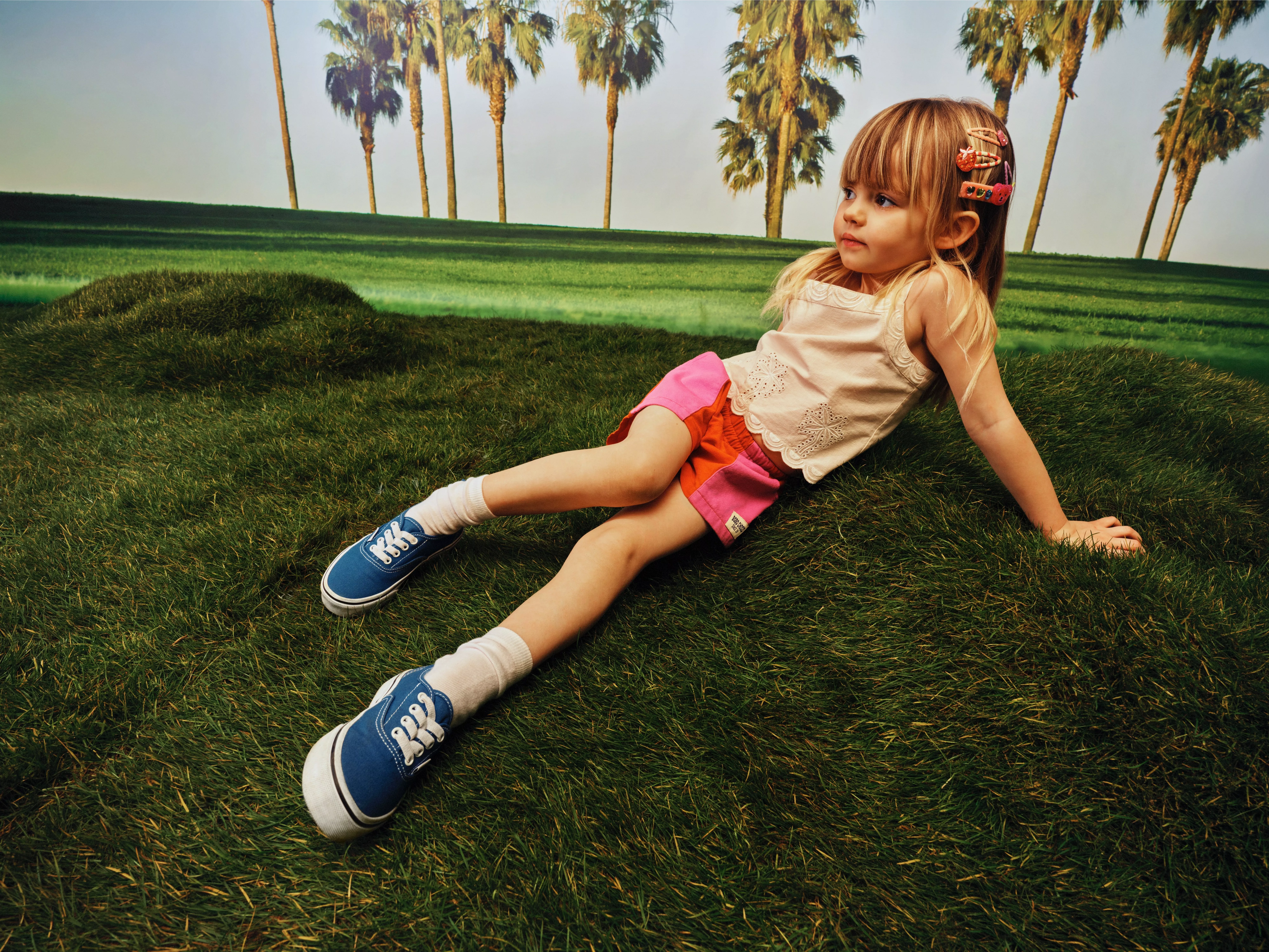 Young girl lounging on green grass in blue trainers, colourful shorts and hair clips, with palm trees behind