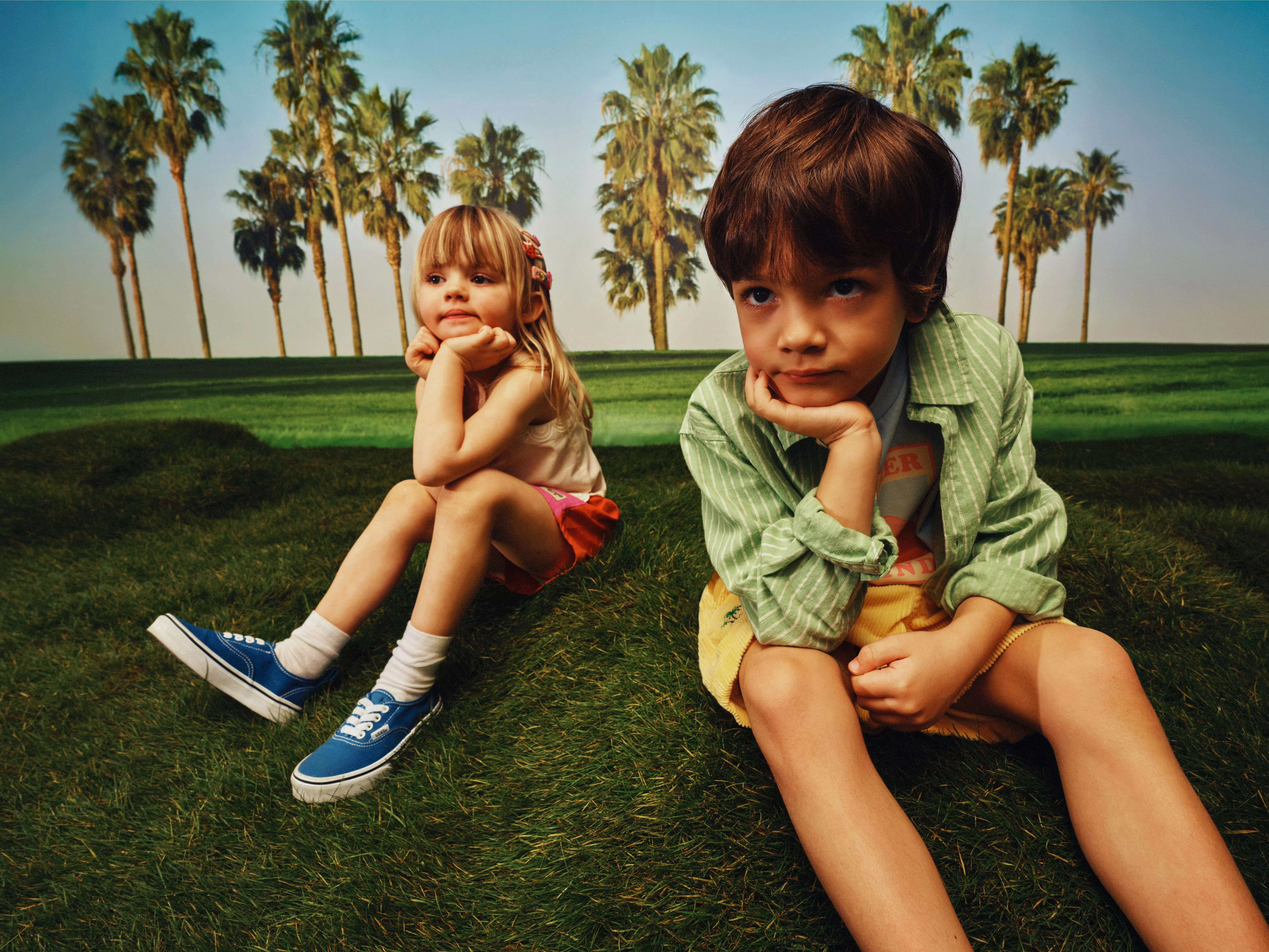 Two children sitting on green grass with palms behind, resting chins on hands in sunny outdoor park scene