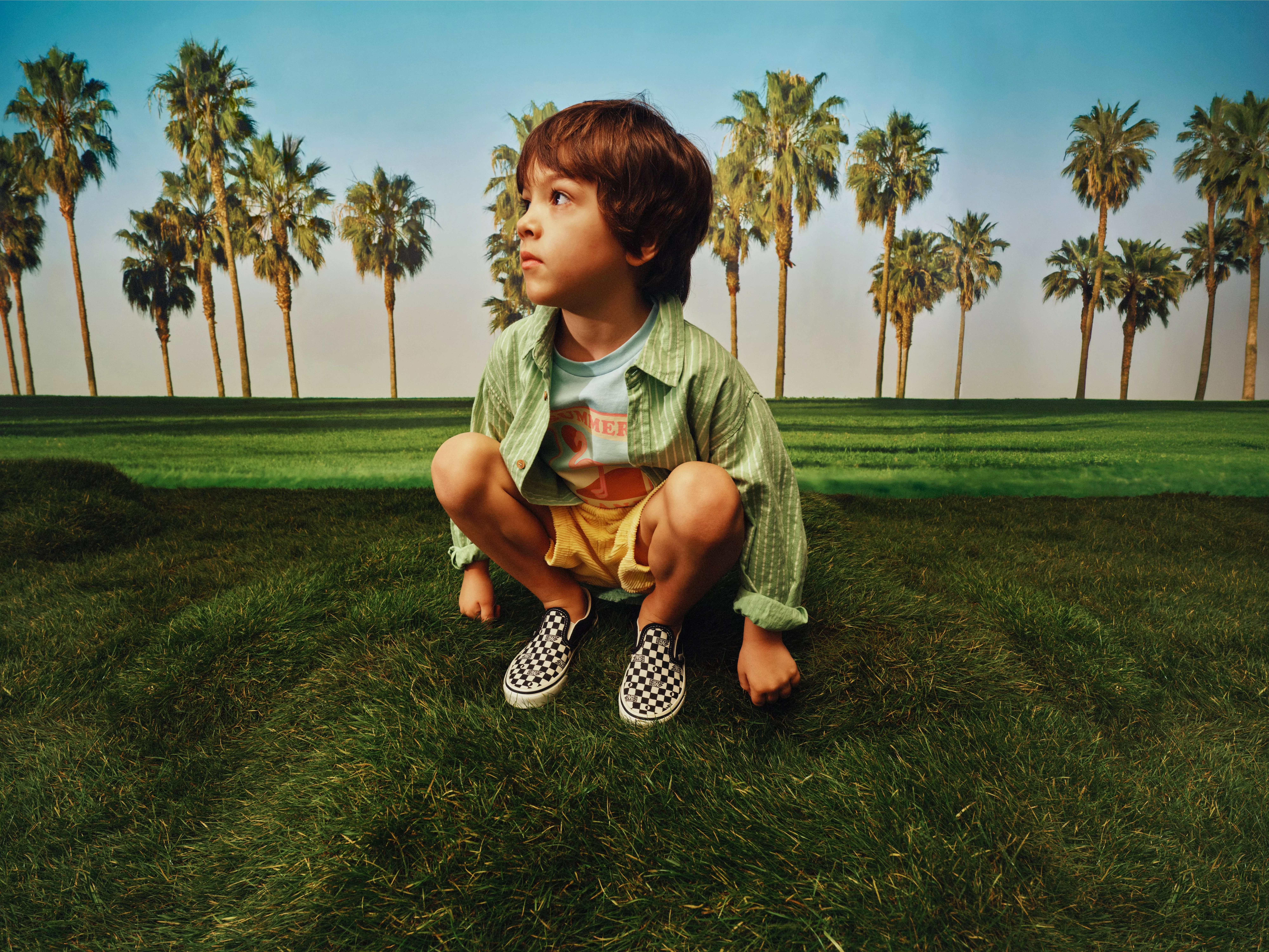 Young boy crouching on green grass in checkerboard slip-ons, with tall palm trees and blue sky backdrop