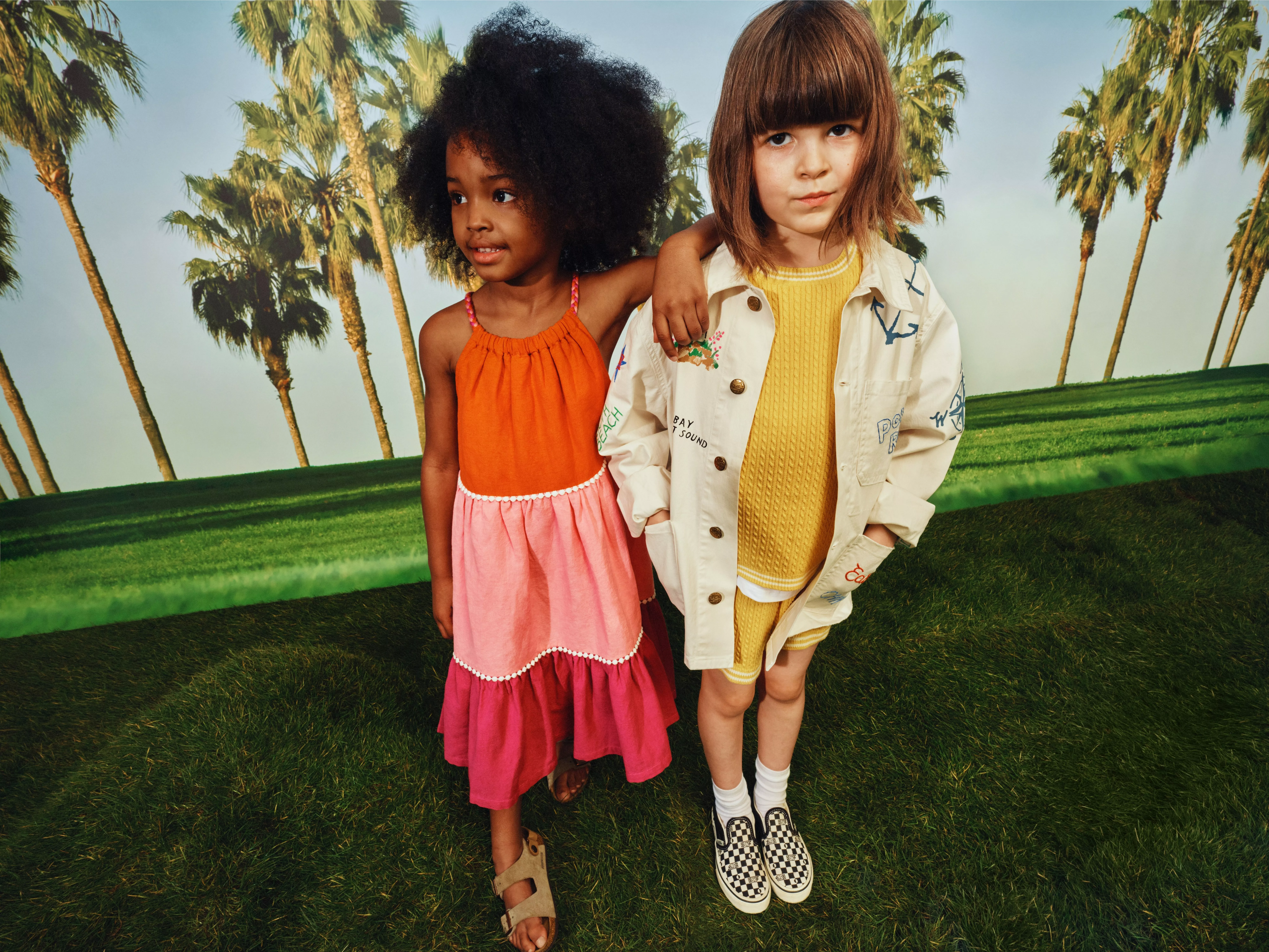 Two children in summer outfits posing on green grass with palm trees, kids fashion photo outdoors.