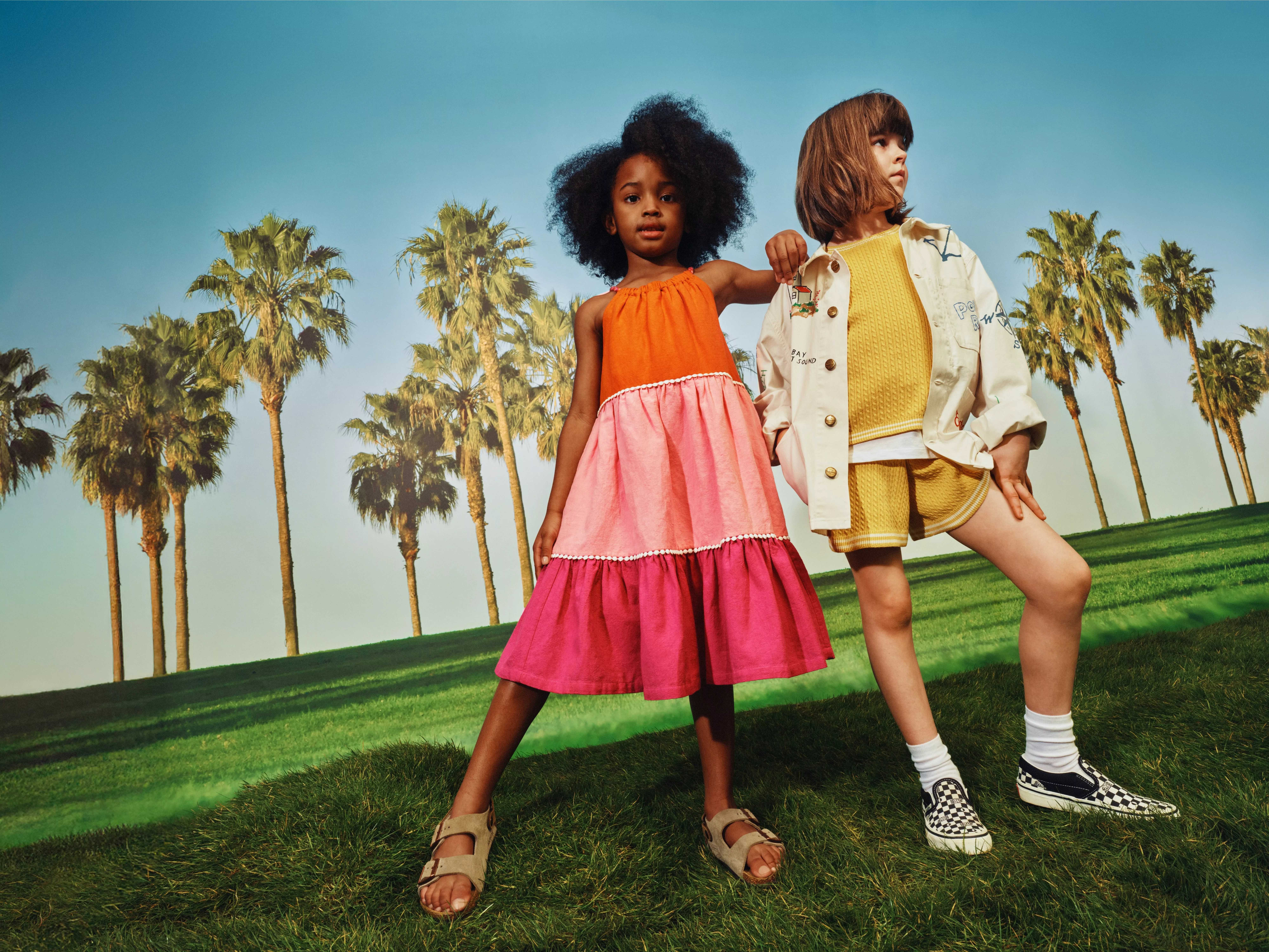Two children modelling colourful summer outfits on a sunny grassy hill with palm trees and blue sky backdrop