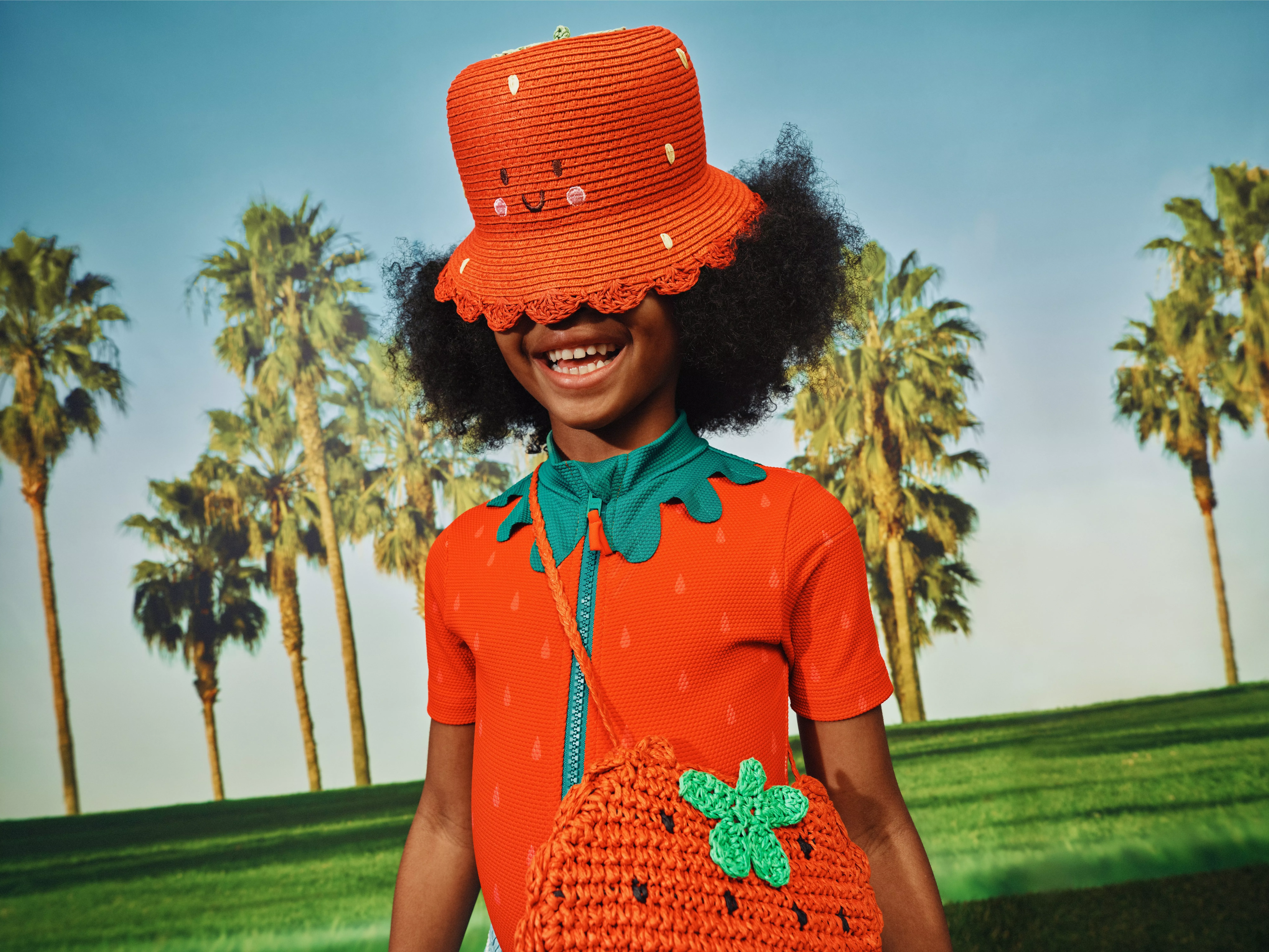 Smiling child in strawberry outfit with orange bucket hat and crocheted bag, outdoors with palm trees in sunshine
