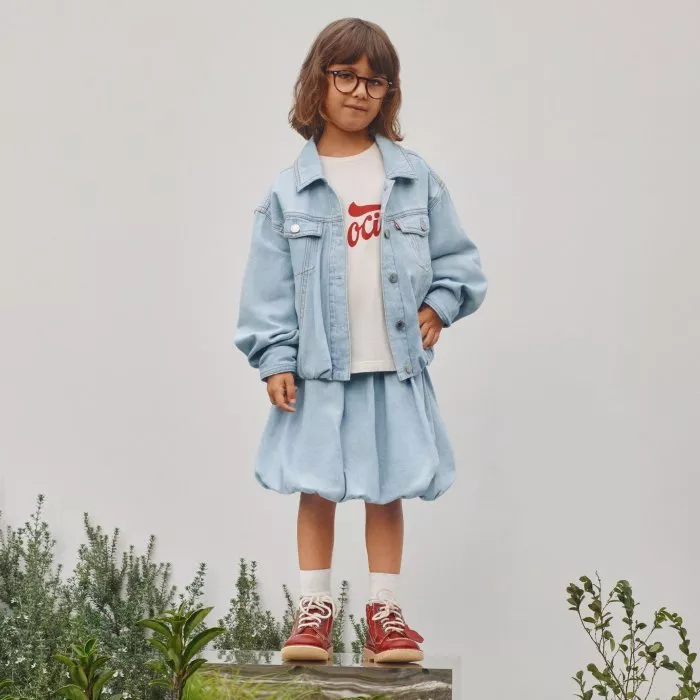 Girl in denim jacket and skirt with glasses and red trainers, standing outdoors against a plain wall