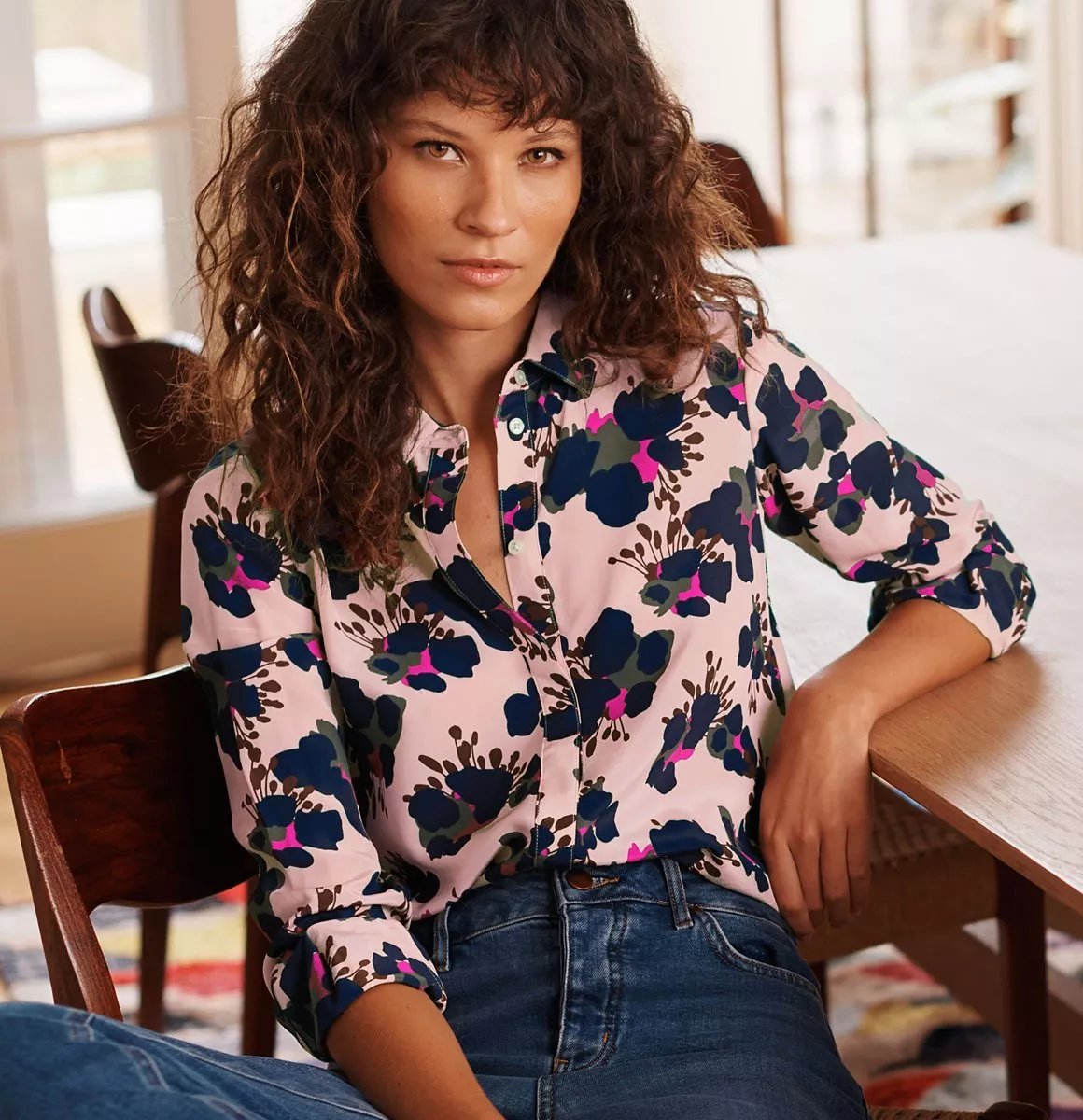 Woman in pink floral blouse and blue jeans, seated at wooden table in bright modern home interior