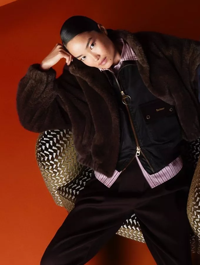 Fashion portrait of a woman in faux fur coat and bomber jacket, reclining in a patterned chair on orange backdrop