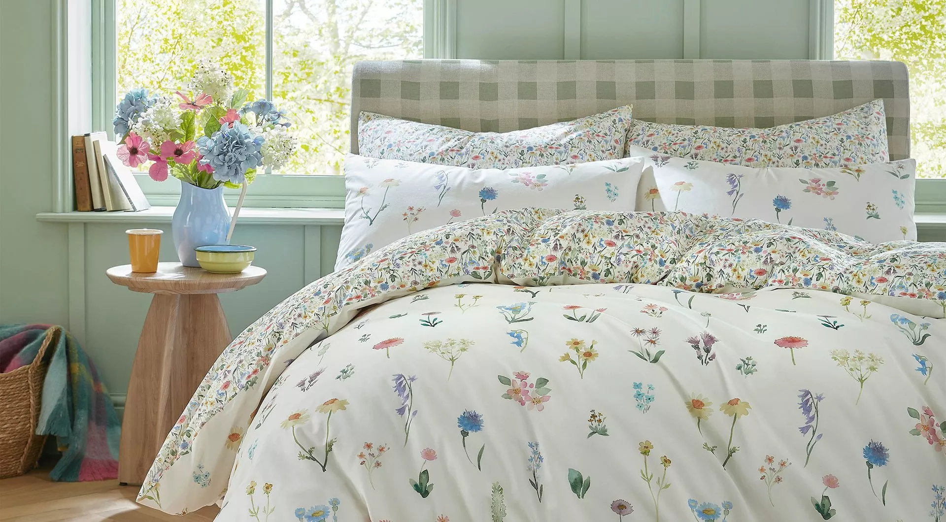 A cosy bed dressed in white bedding with a colourful delicate wildflower print, set in a light green bedroom with fresh flowers in a blue vase on a side table.