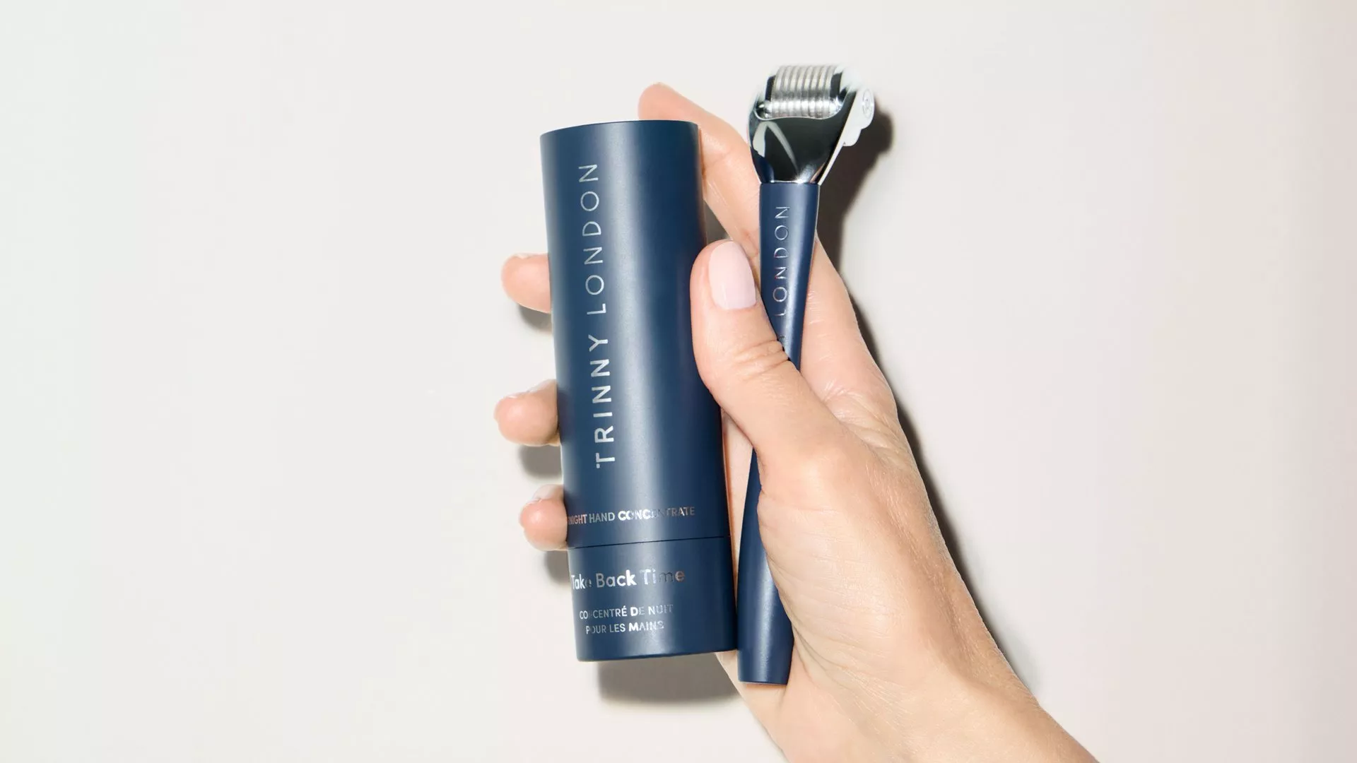 A hand holding a navy blue Trinny London Take Back Time skincare tube alongside a matching micro-needle roller tool against a light neutral background.