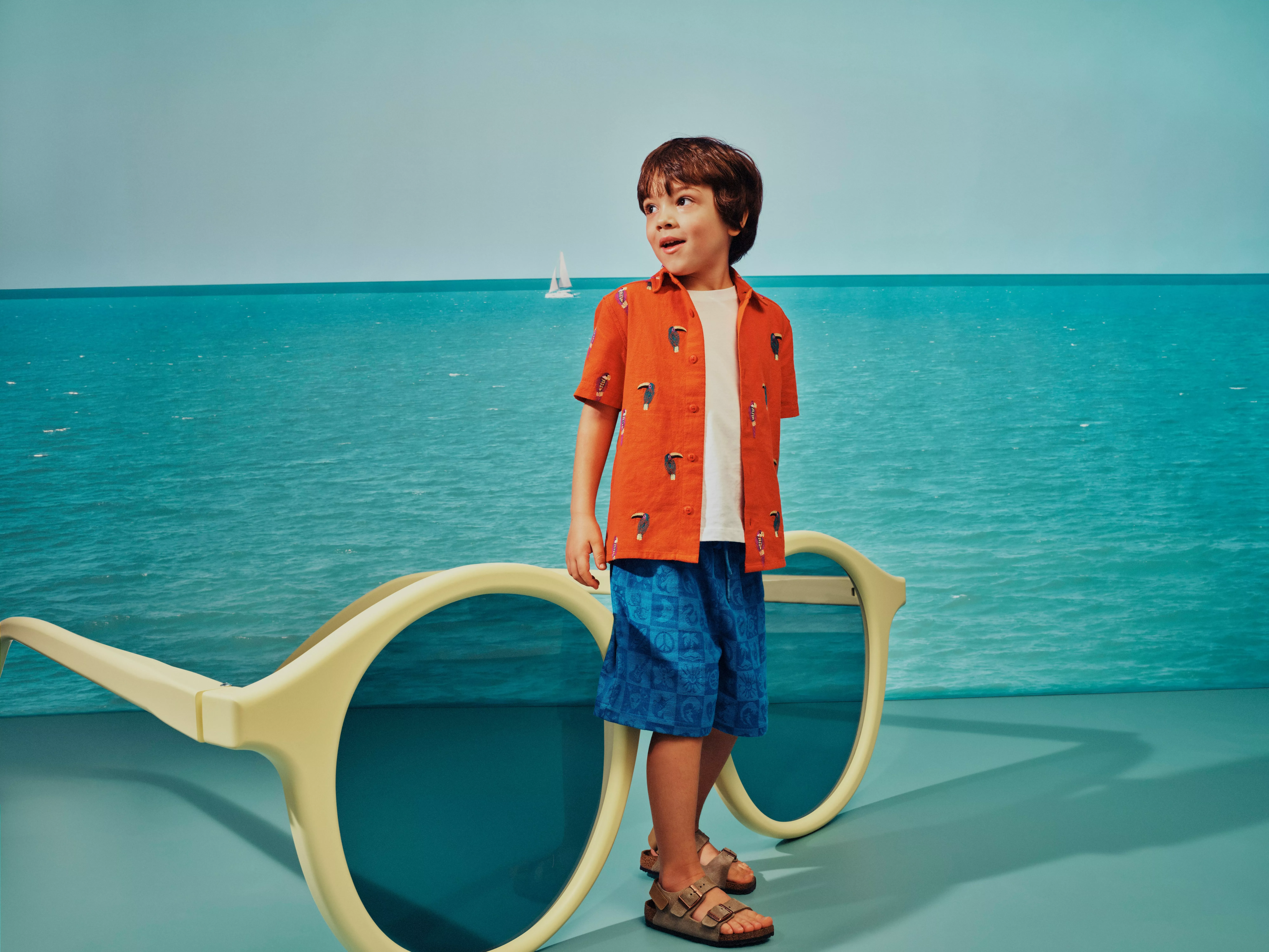 Boy in orange shirt and blue shorts standing by oversized sunglasses, with calm sea and sailboat backdrop