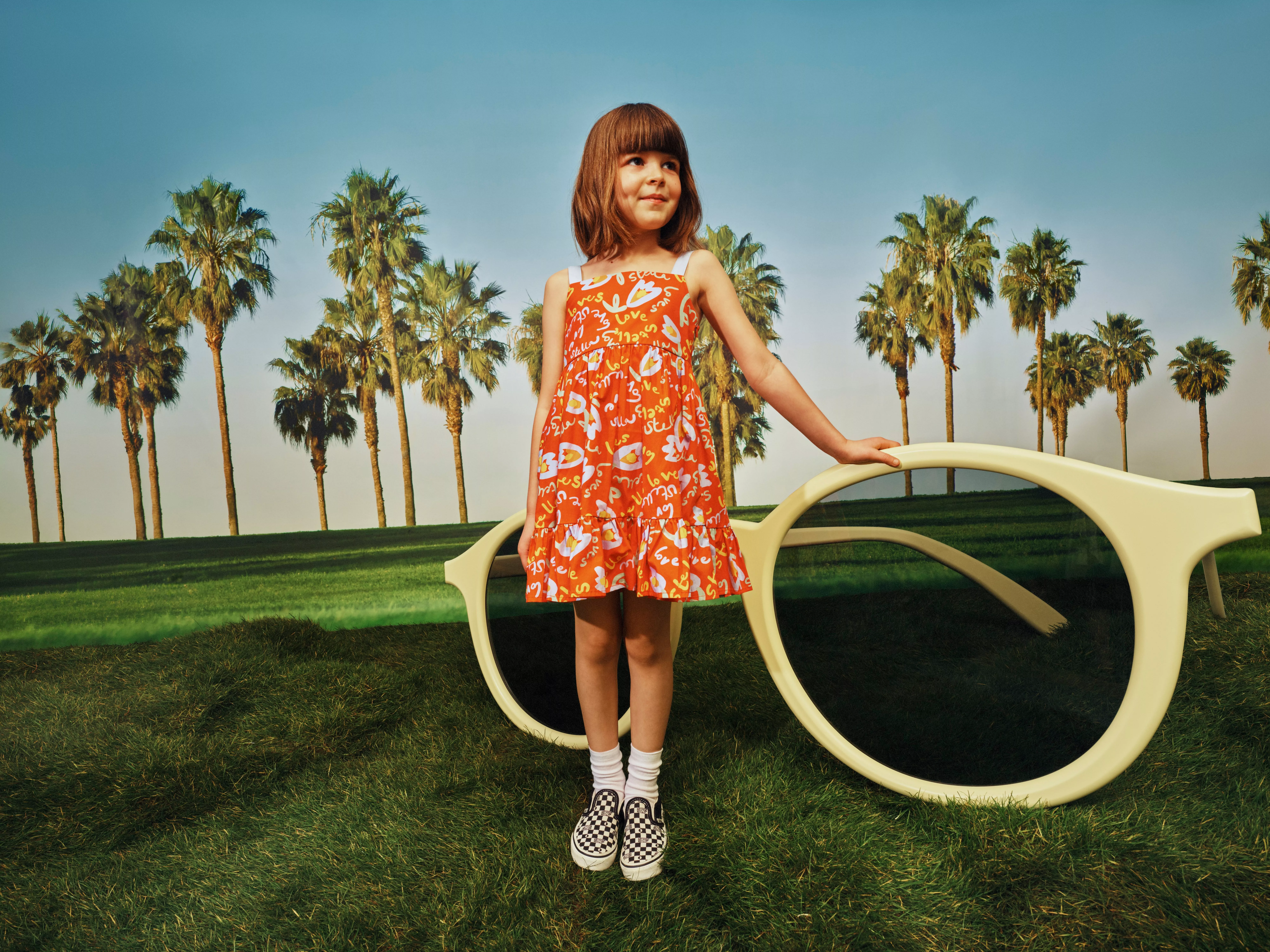 Smiling child in denim outfit sitting on a yellow lounge chair on green lawn with palm trees outdoors
