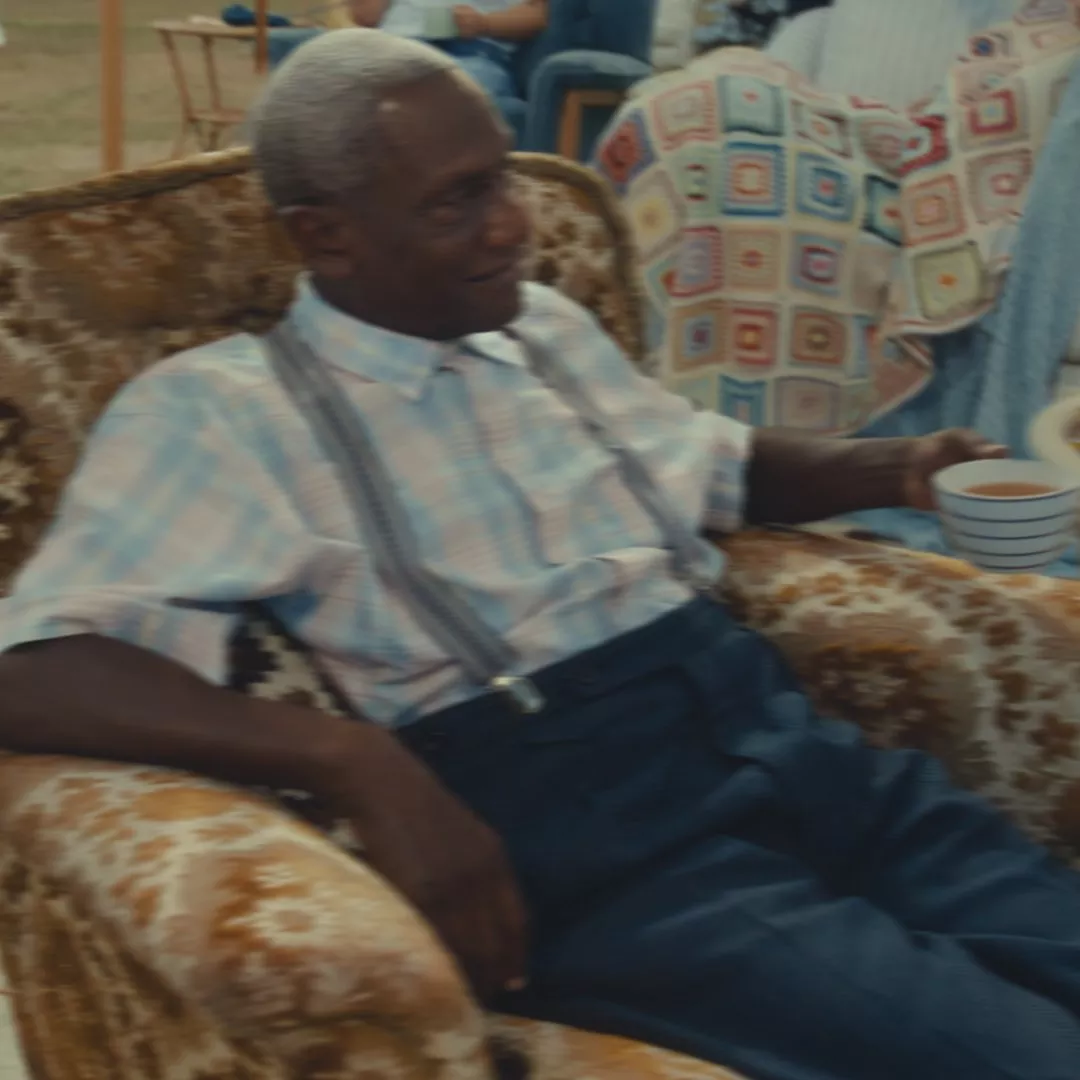Elderly man with grey hair in a checkered shirt and suspenders, sits on a patterned chair holding a cup.